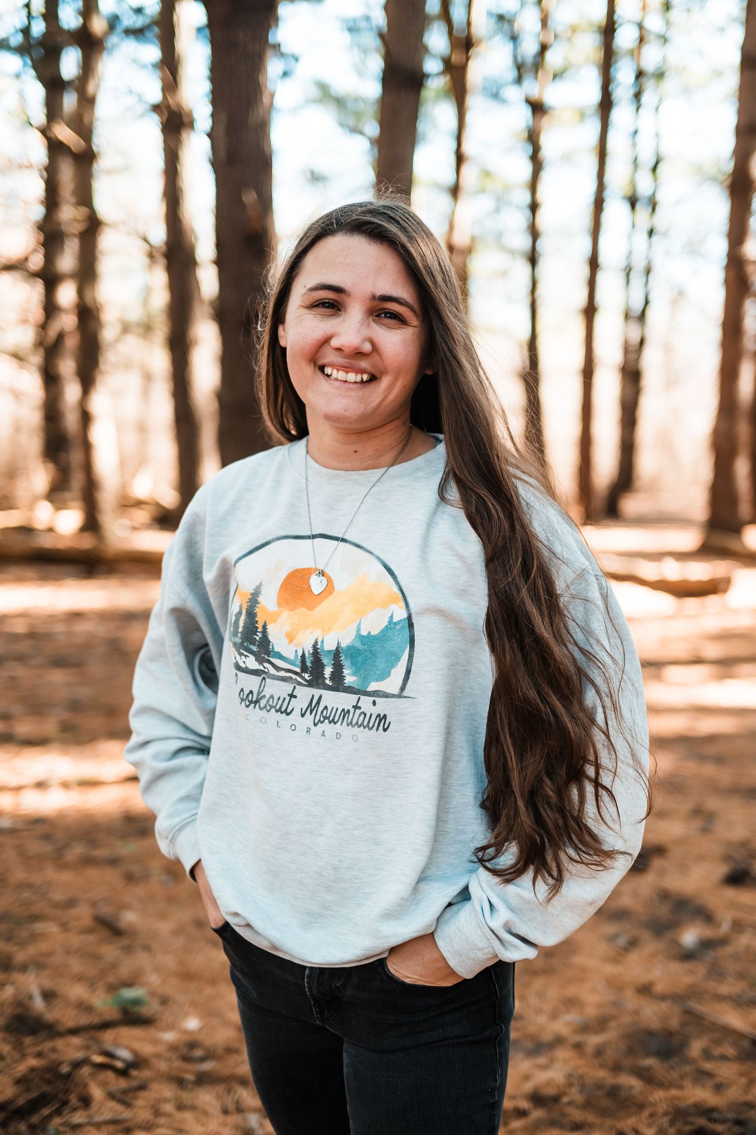 A person wearing a gray sweatshirt with an outdoor adventure graphic design stands smiling in a sunlit forest.
