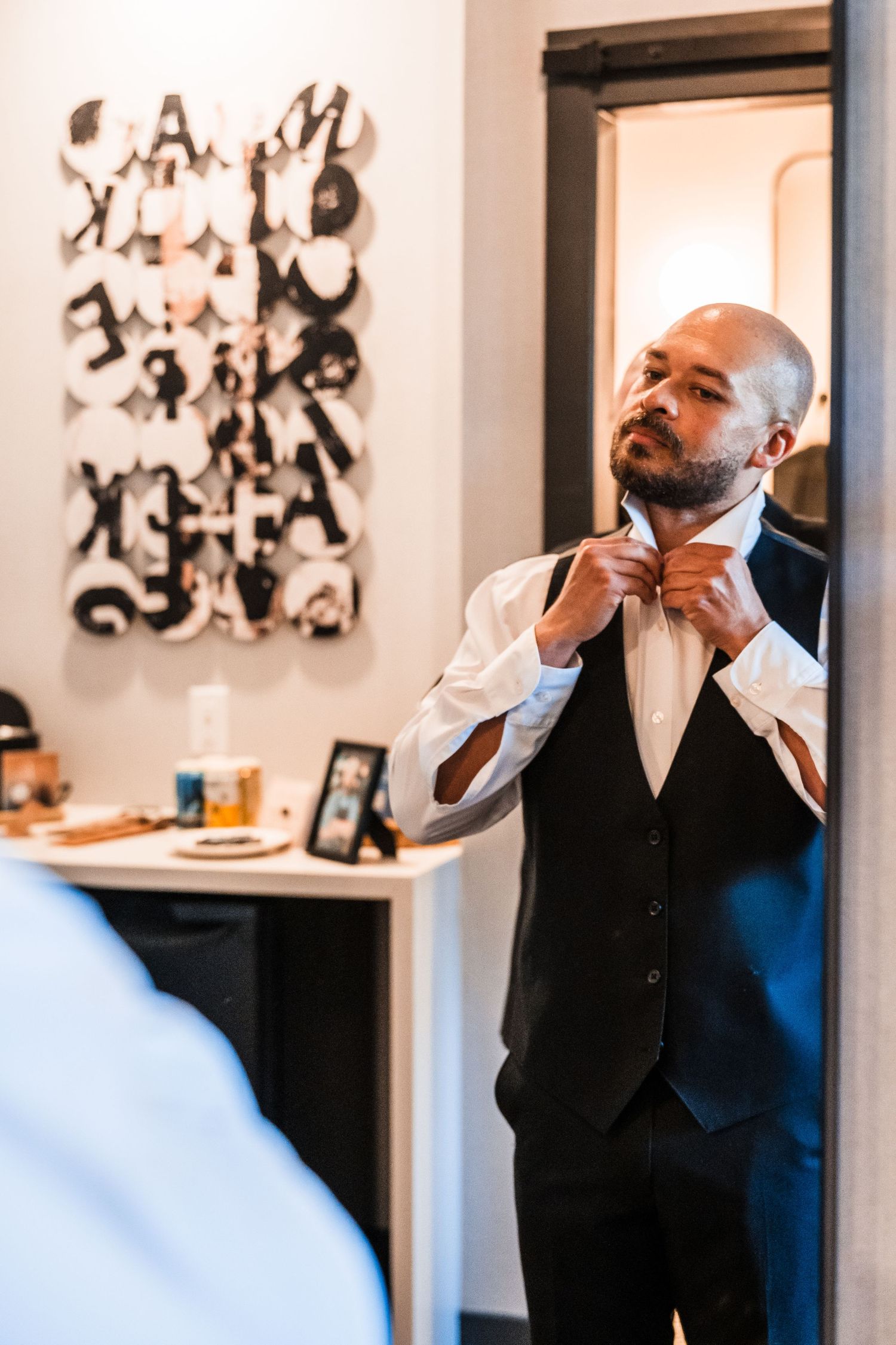 Groom in black vest adjusts bow tie while looking in mirror before wedding ceremony.