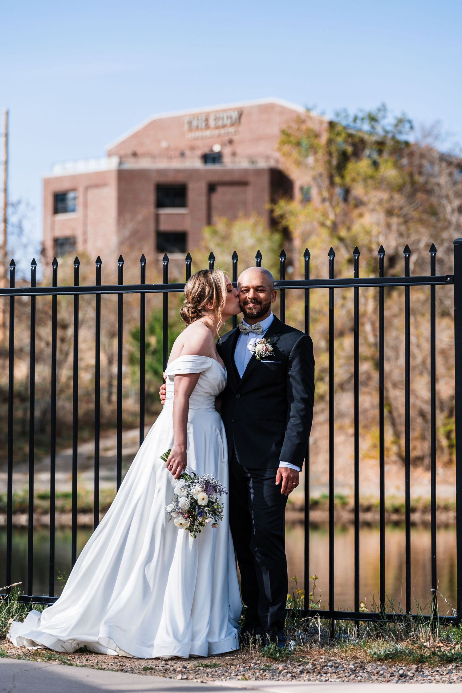 Bride kissing groom on cheek while posing by black iron fence with brick building backdrop on wedding day.