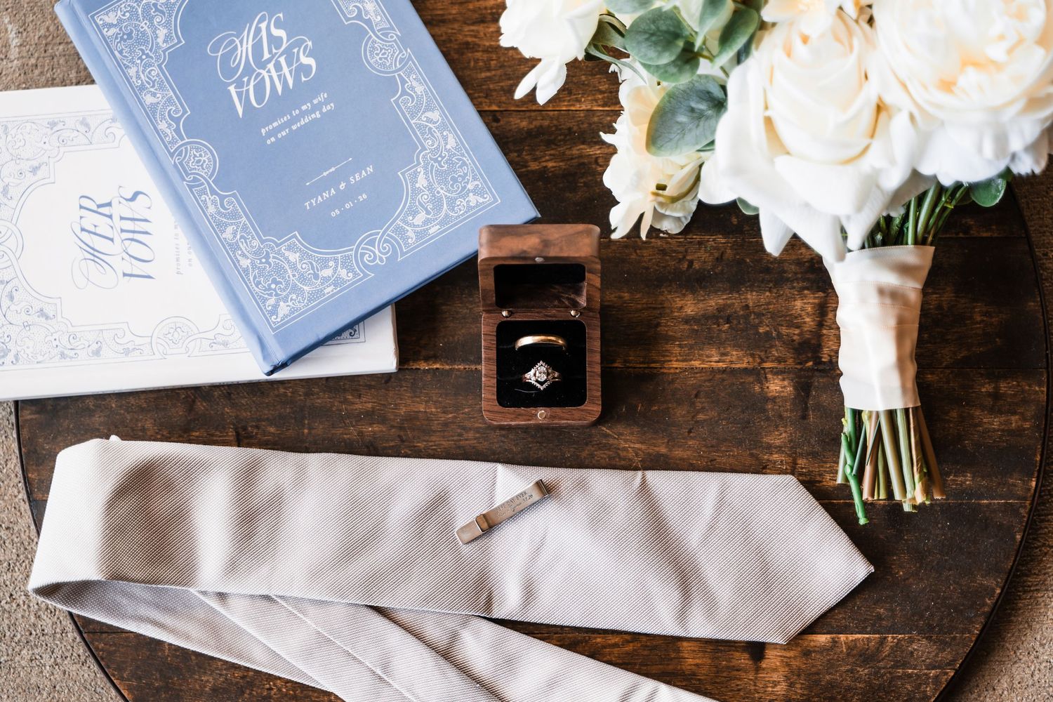 Wedding flat lay with vow books, ring box, diamond ring, silk tie, cufflink, and white rose bouquet on wood table.