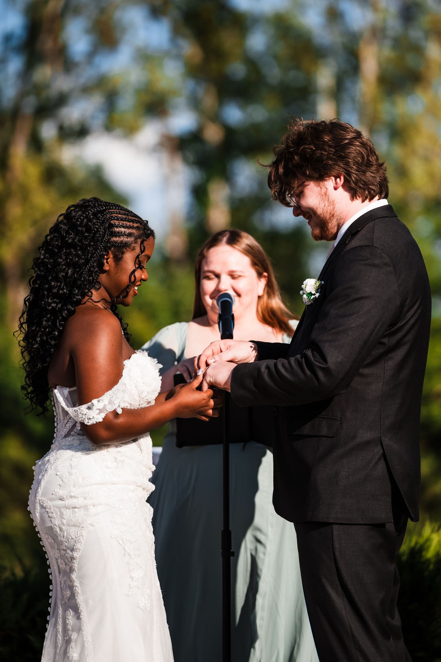 Bride placing wedding ring on grooms finger during outdoor ceremony with officiant in background.