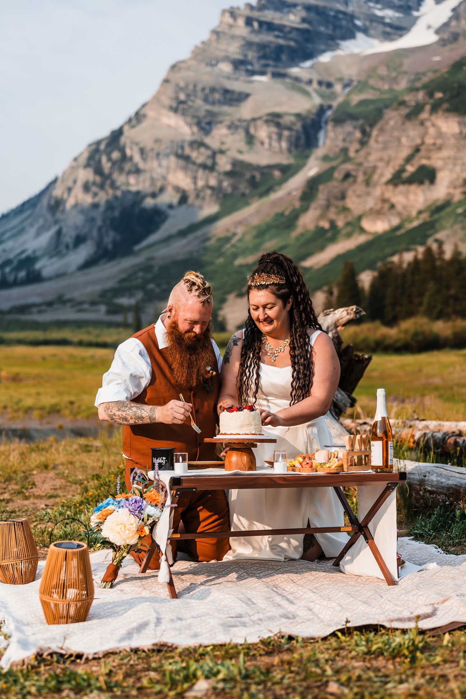 Couple cuts wedding cake at rustic outdoor picnic elopement with mountain backdrop in Colorado.