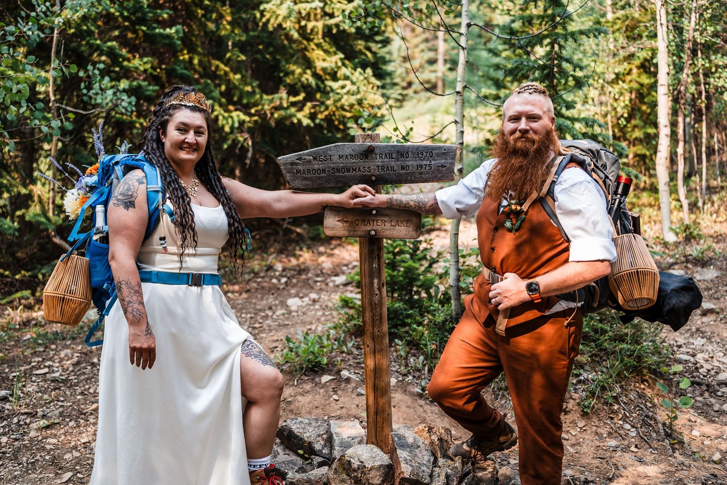 Bride and groom in hiking gear pose at a forest trail sign during their outdoor mountain elopement.