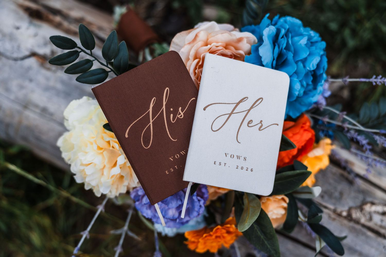 His and Hers wedding vow books on colorful bridal bouquet with blue, orange, and white flowers.
