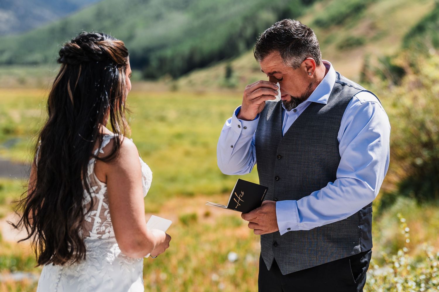 Emotional groom wipes tears while reading heartfelt wedding vows to his bride in a mountain meadow setting.