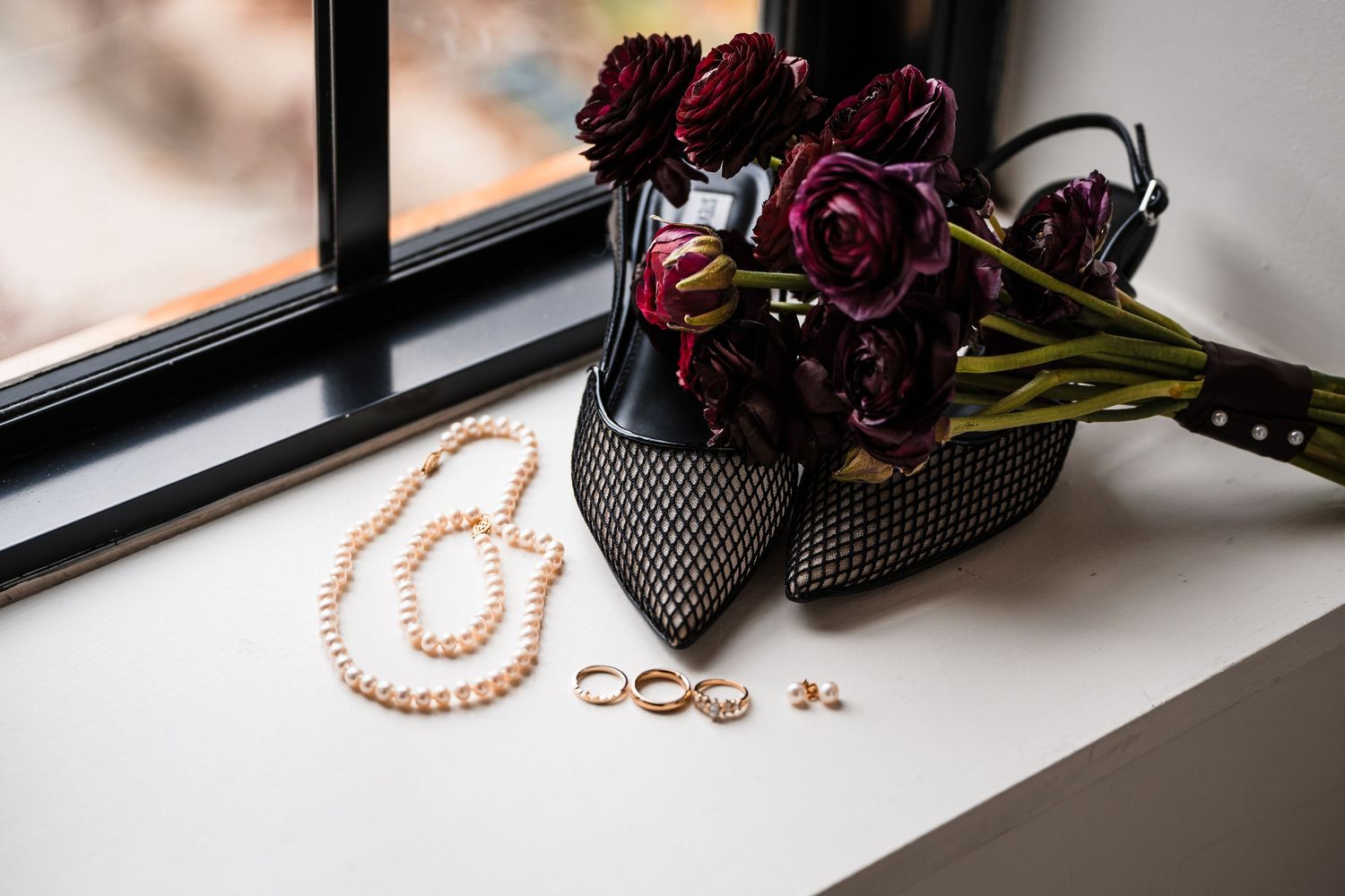 Pink pearl necklace and gold rings arranged on white windowsill with dark flowers and accessories in background.