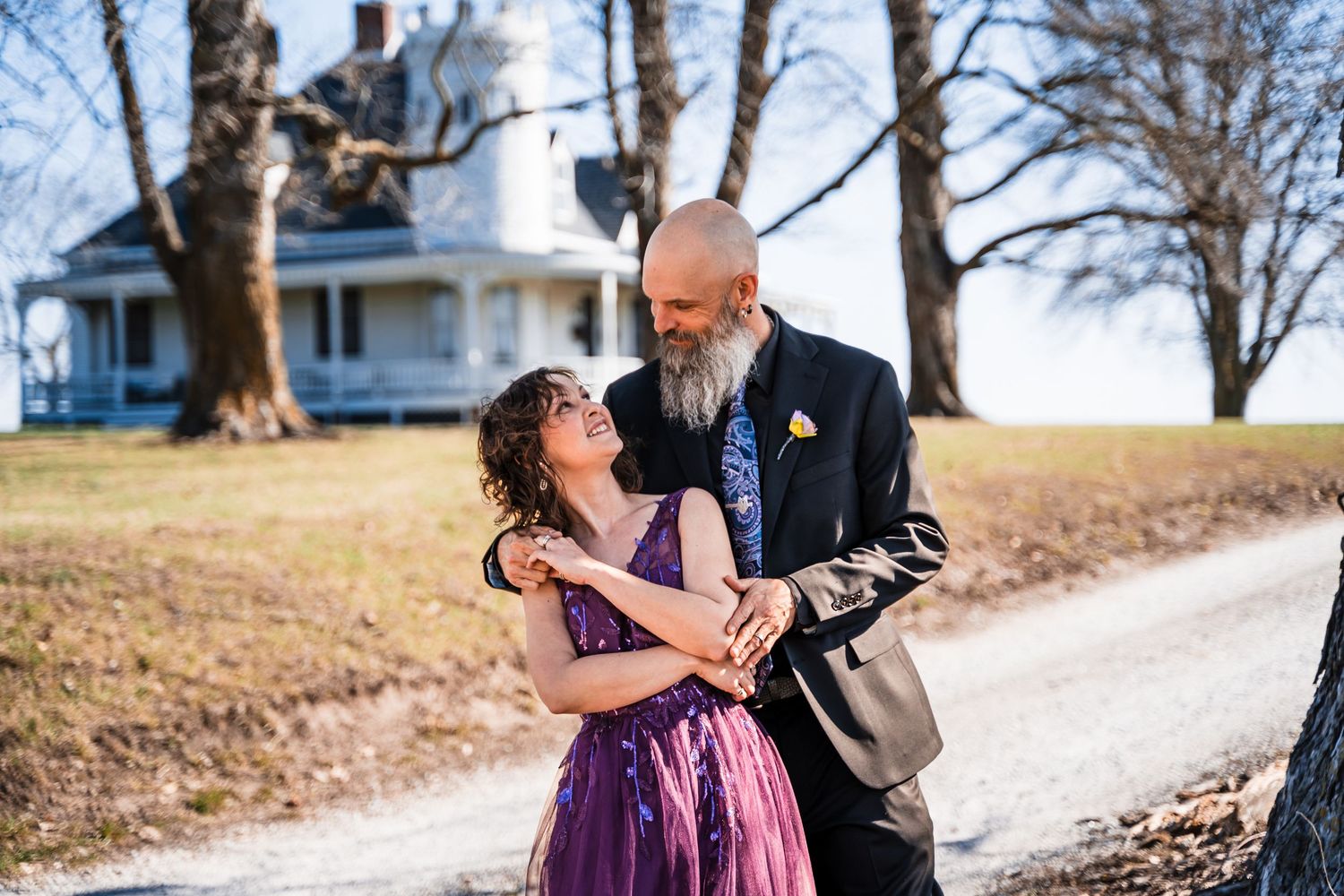 Couple embracing outdoors near a historic white farmhouse, man in suit with beard, woman in purple dress.