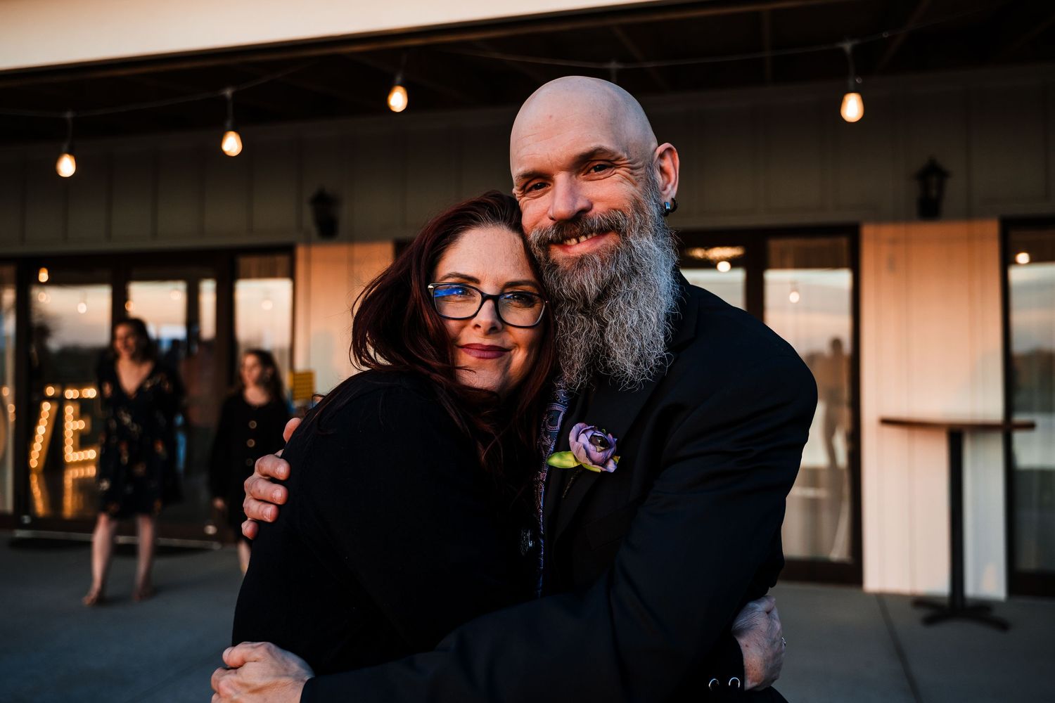 Bald bearded man and woman with glasses sharing warm hug at outdoor evening event.
