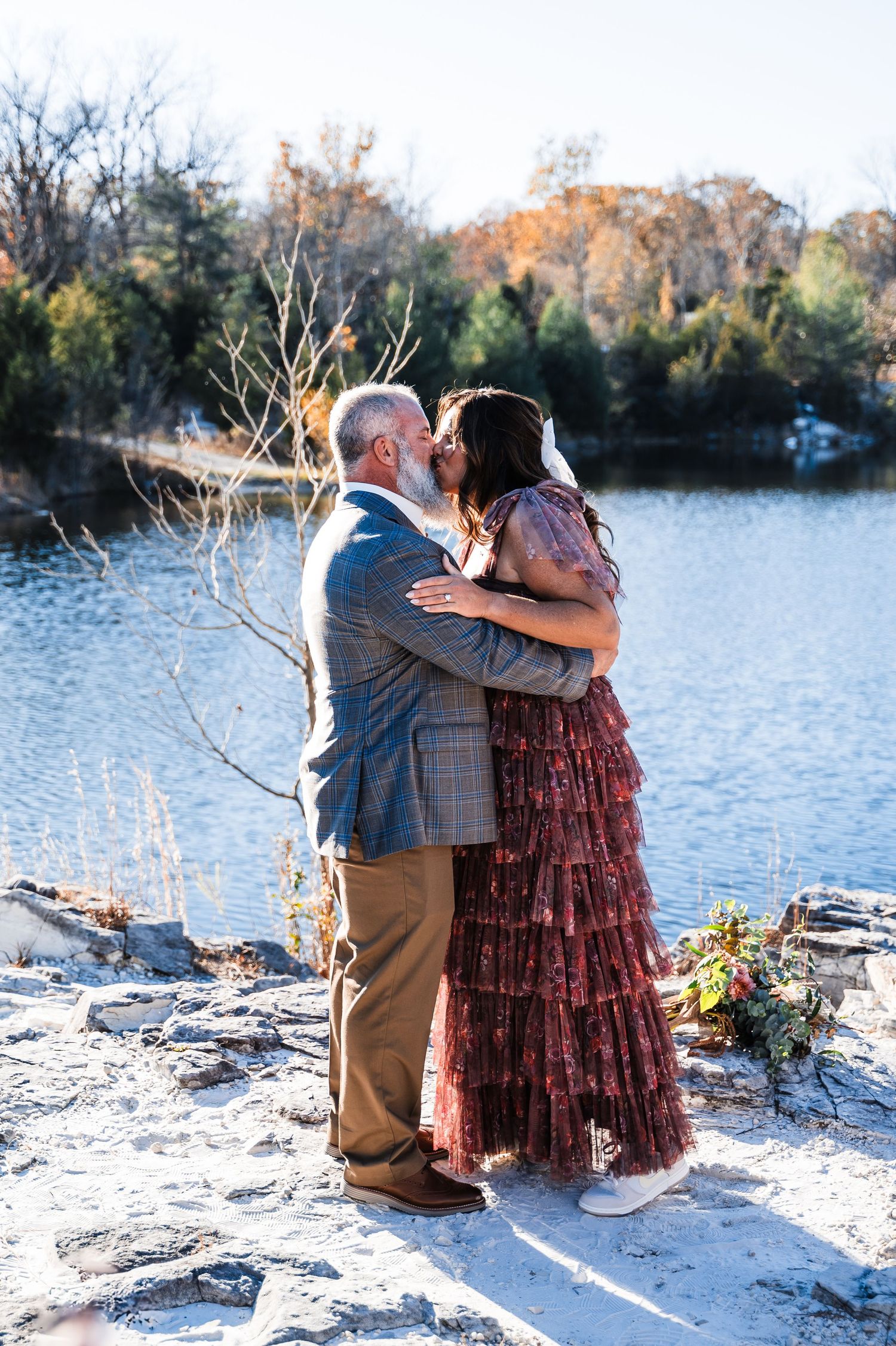A romantic couple embraces by a serene lake on an autumn day with one wearing a floral dress and the other in plaid.