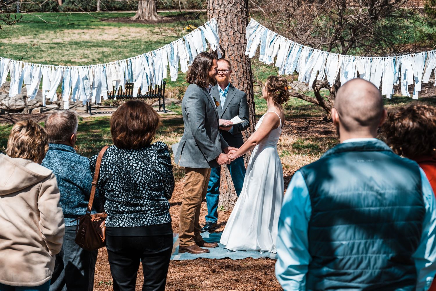 An intimate outdoor wedding ceremony under trees decorated with white fabric streamers.