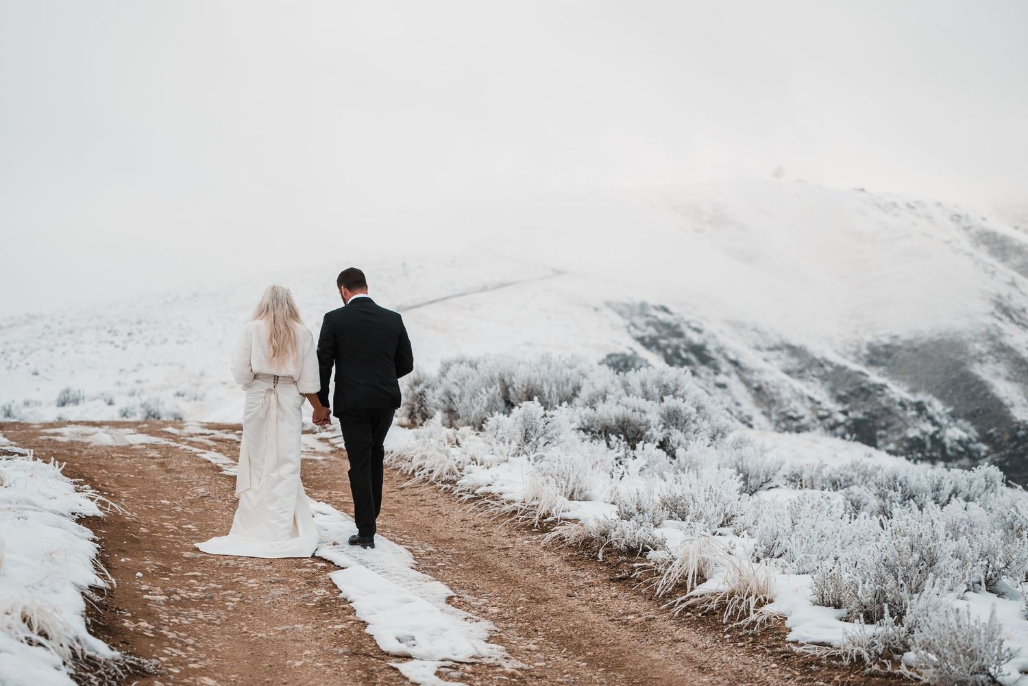 Newlyweds walking along snowy mountain path with dramatic views.