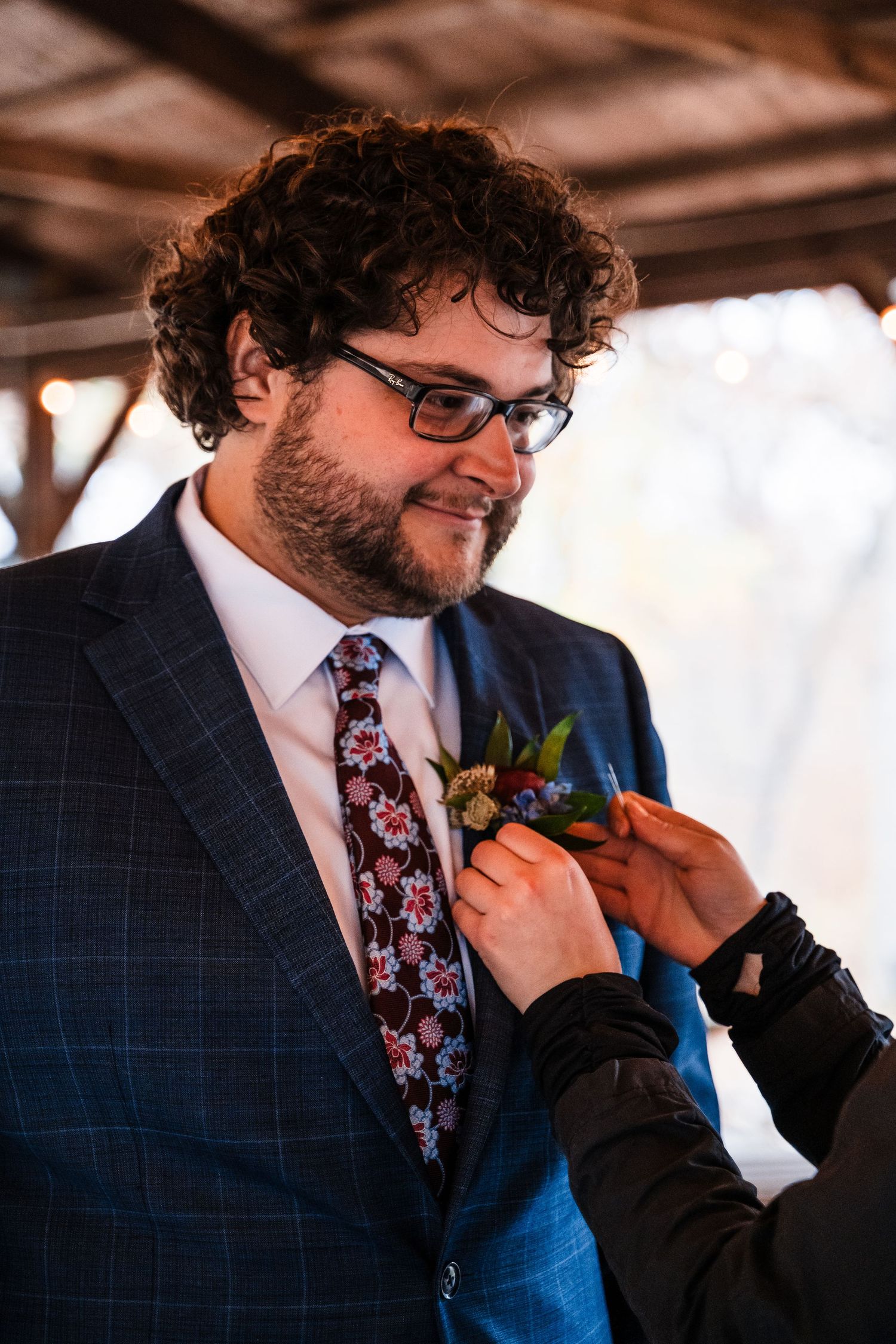 Groom in navy suit having boutonniere pinned on at rustic wedding venue.