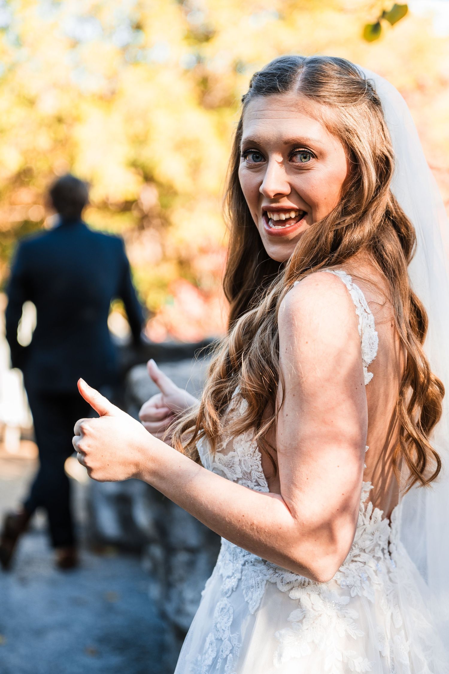 Bride in a white lace dress giving thumbs up with autumn foliage in background.