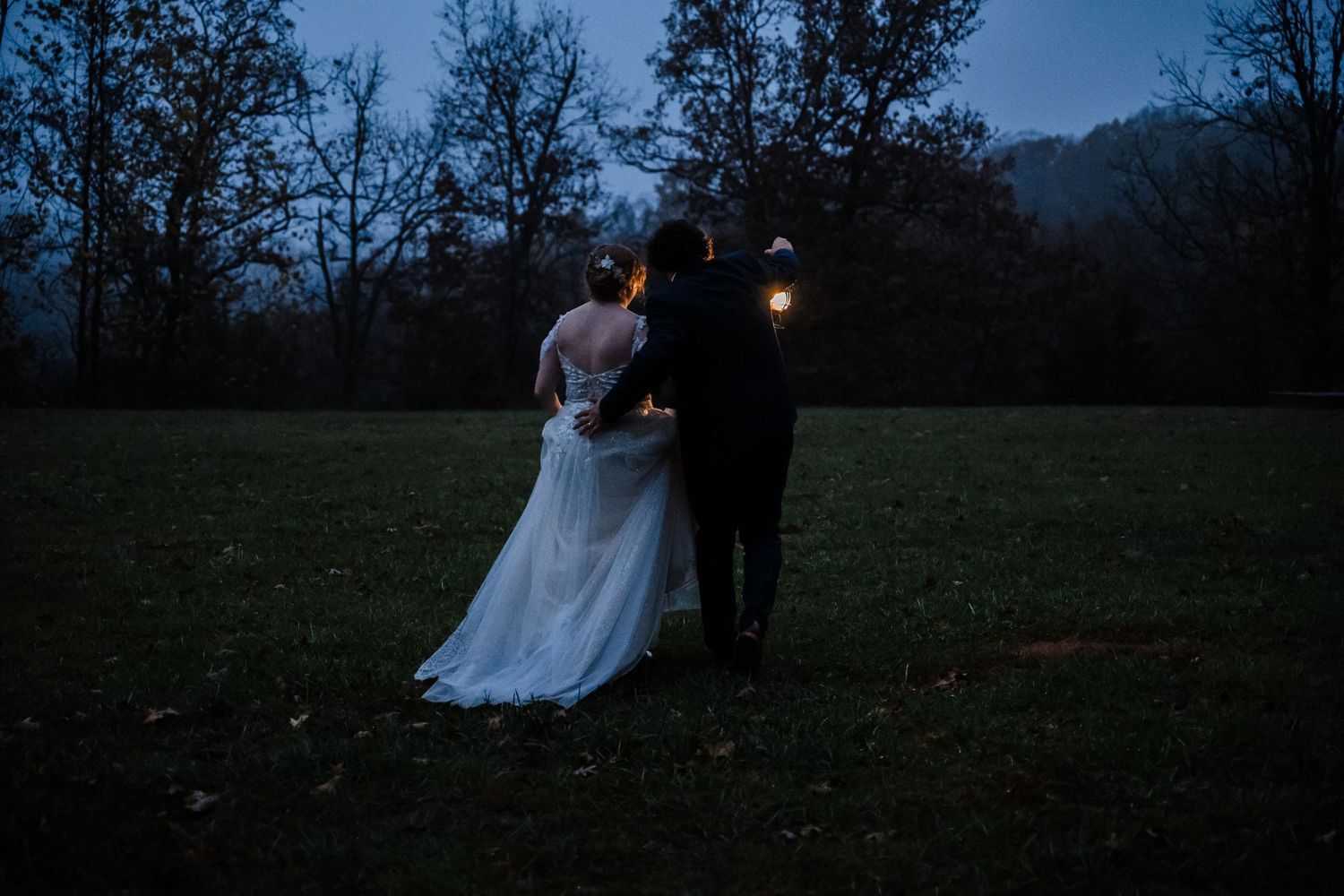 A couple stands together at dusk in a field with glowing lights behind them.
