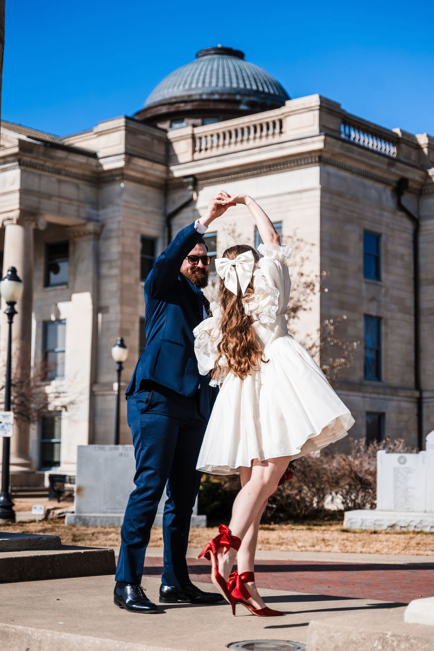 Couple dances outside historic domed building, bride wearing short dress and red heels.