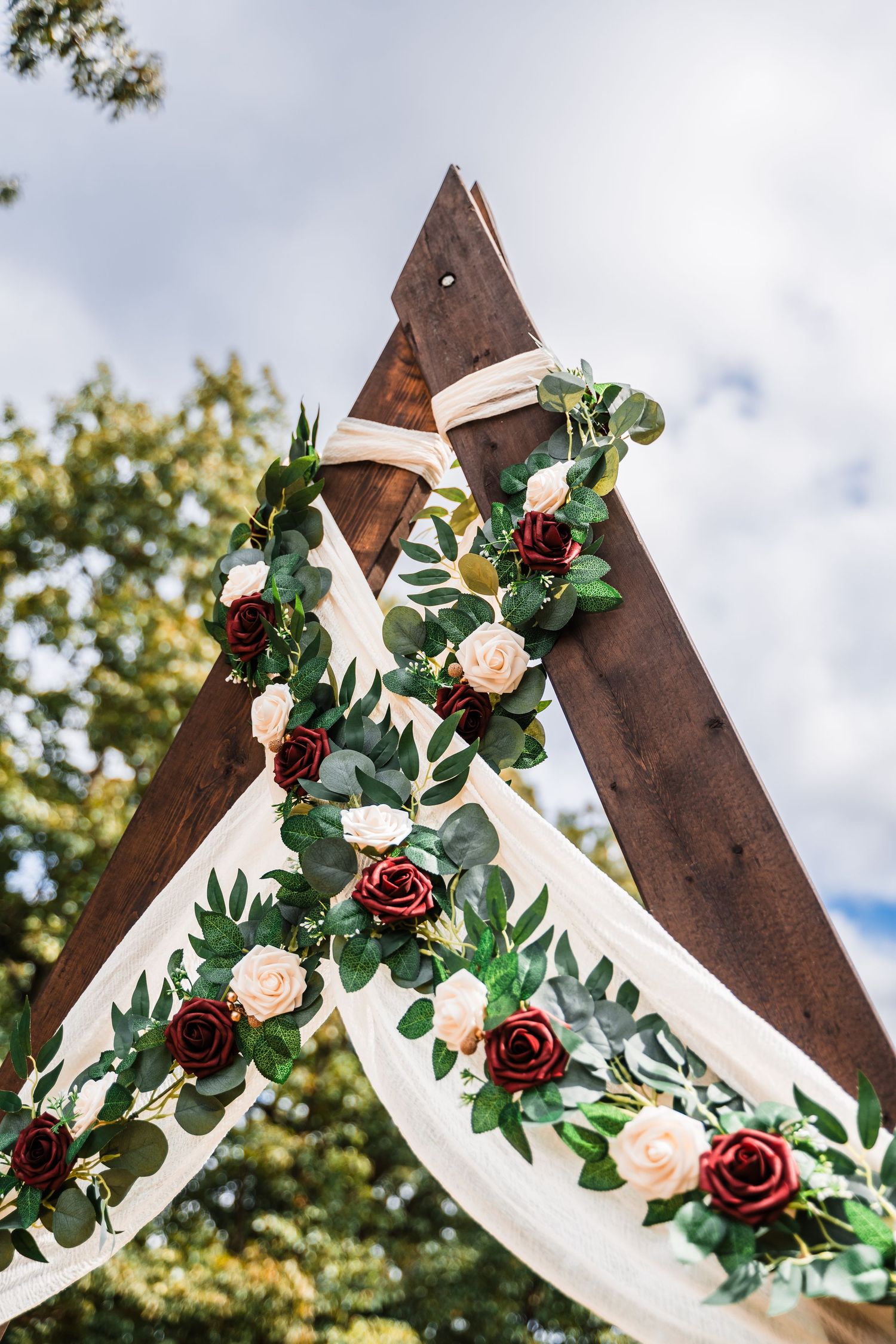 A rustic wooden wedding arch decorated with burgundy and blush roses, white fabric draping, and eucalyptus greenery.