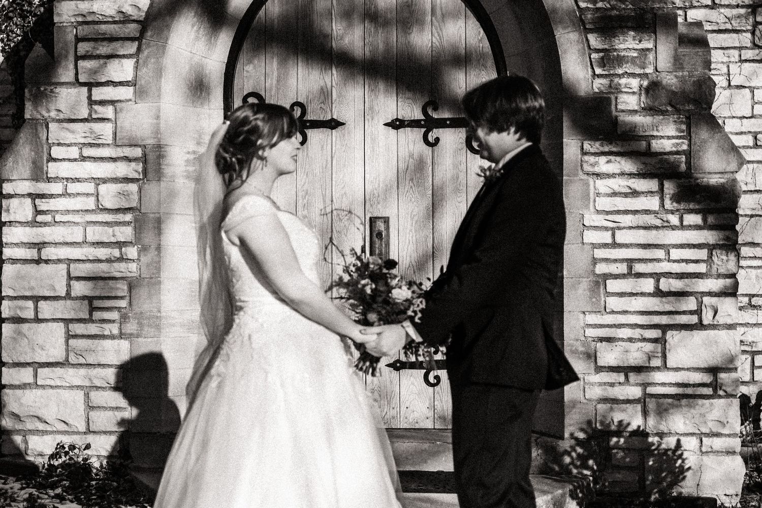 Black and white wedding photo of a couple exchanging vows under a brick archway.
