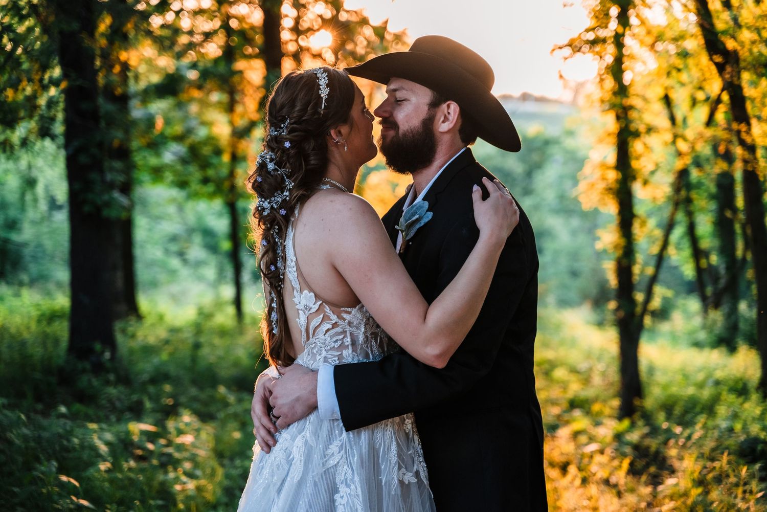 A romantic moment between a couple in formal attire standing in a sunlit forest at golden hour.