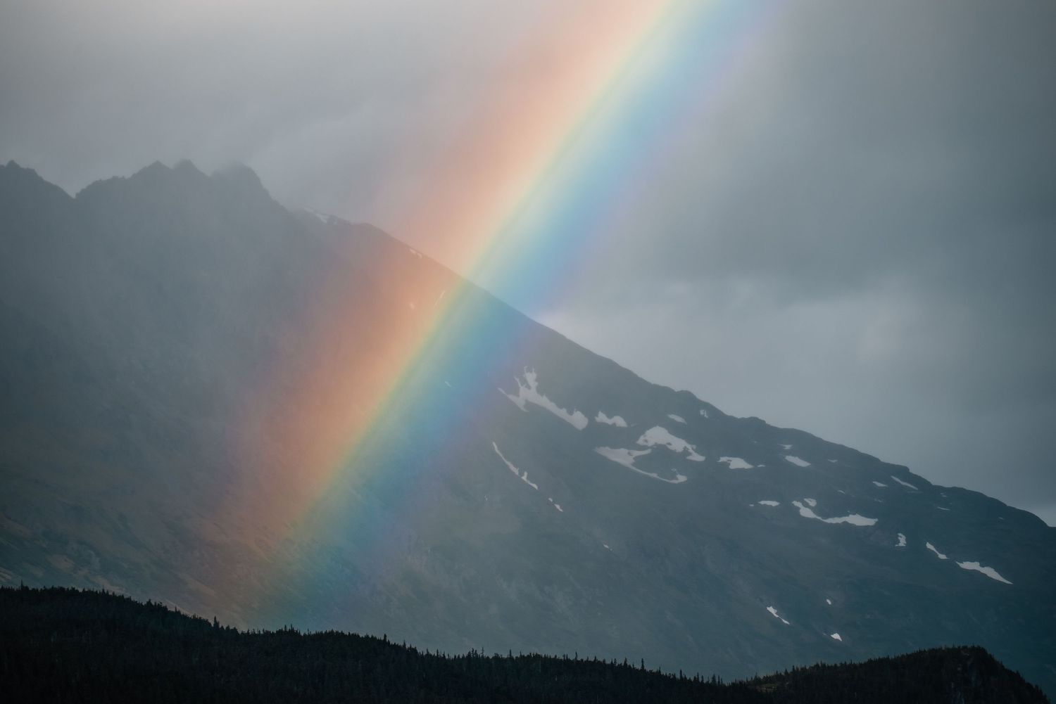 A vibrant rainbow arches over misty mountain peaks with patches of snow and a dark forest silhouette below.