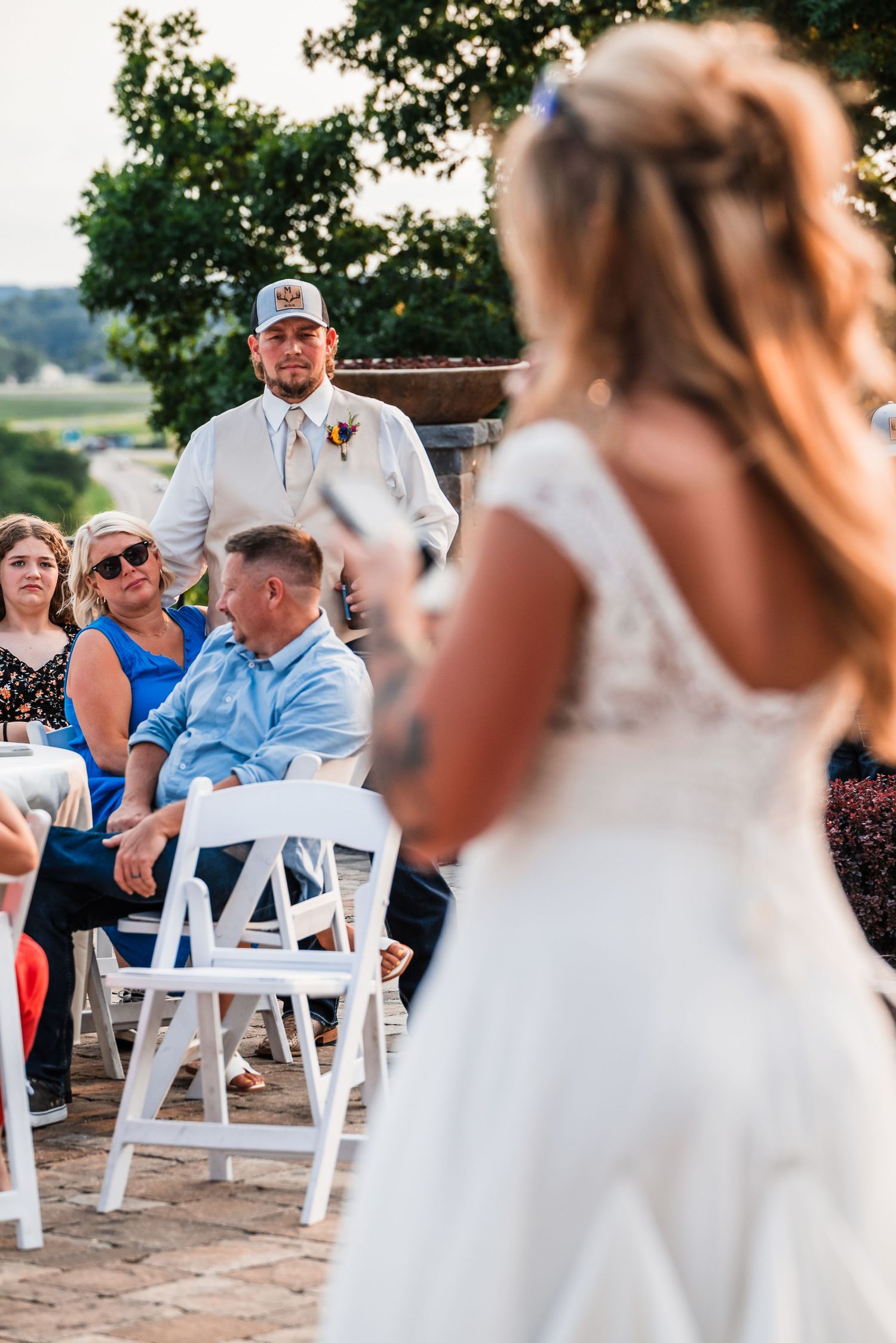 Wedding guests seated in white chairs at an outdoor ceremony with brick backdrop.