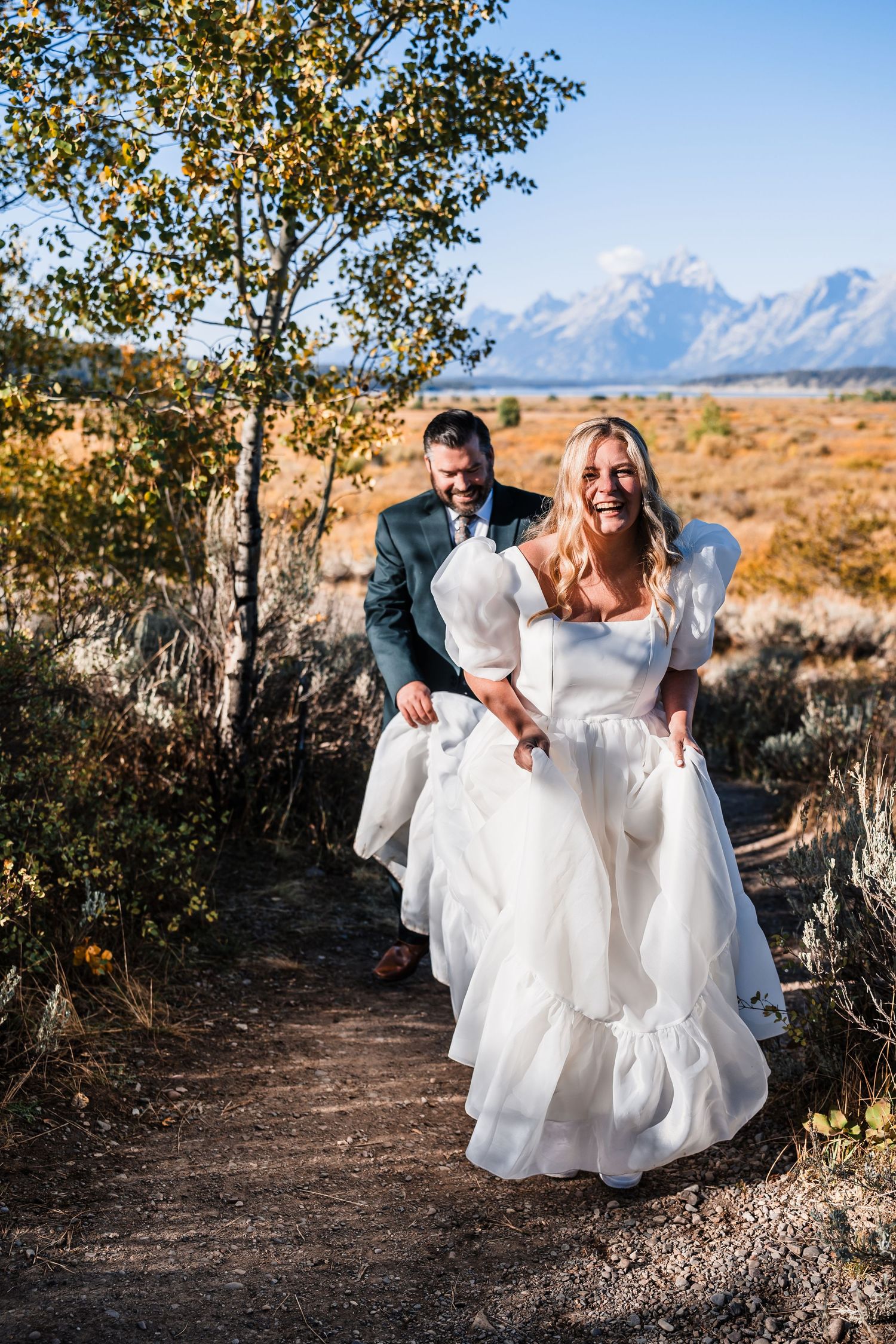 A couple dances playfully on a dirt path with autumn trees and mountains in the background.
