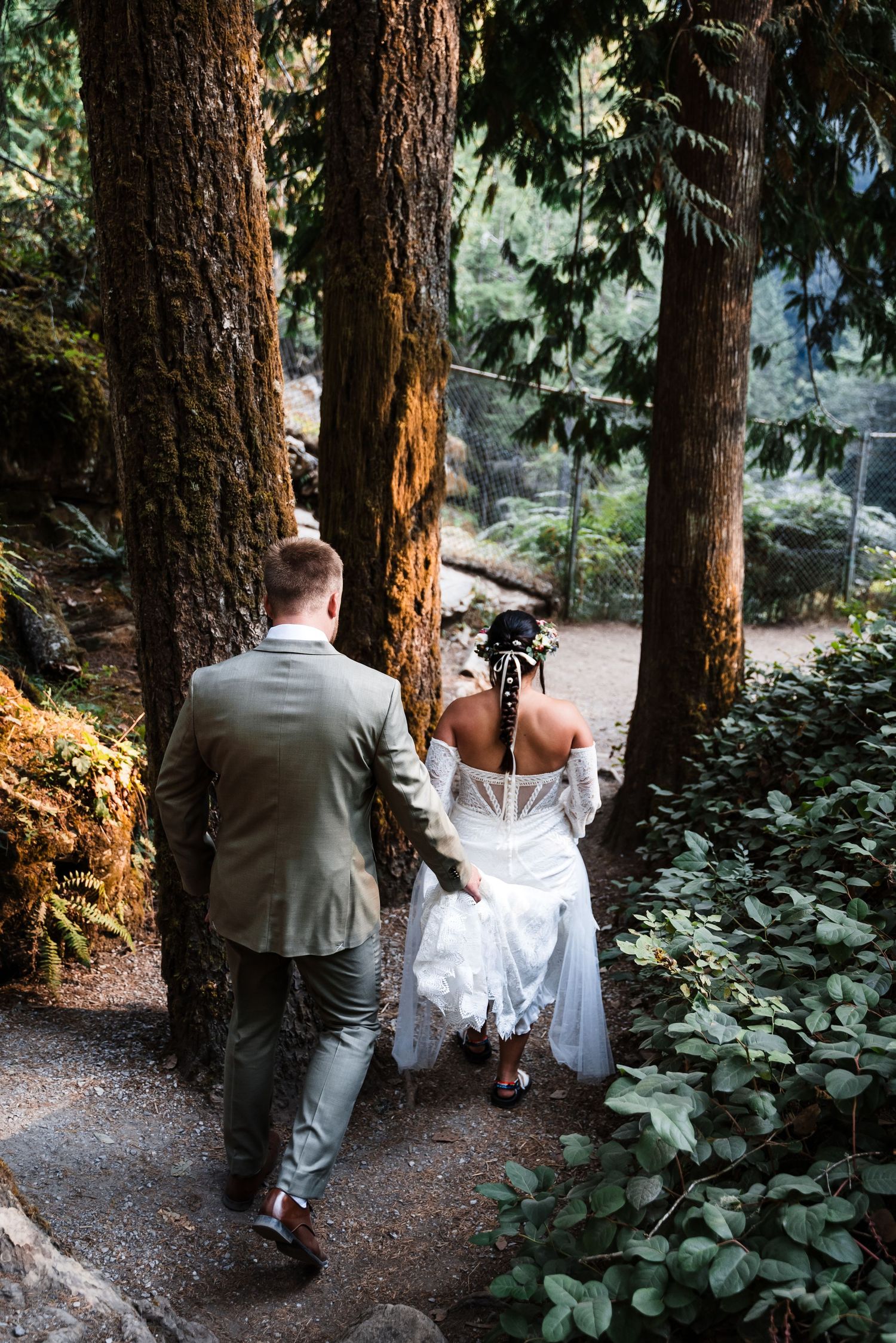 A couple in wedding attire walking down a forest path surrounded by tall pine trees and lush greenery.