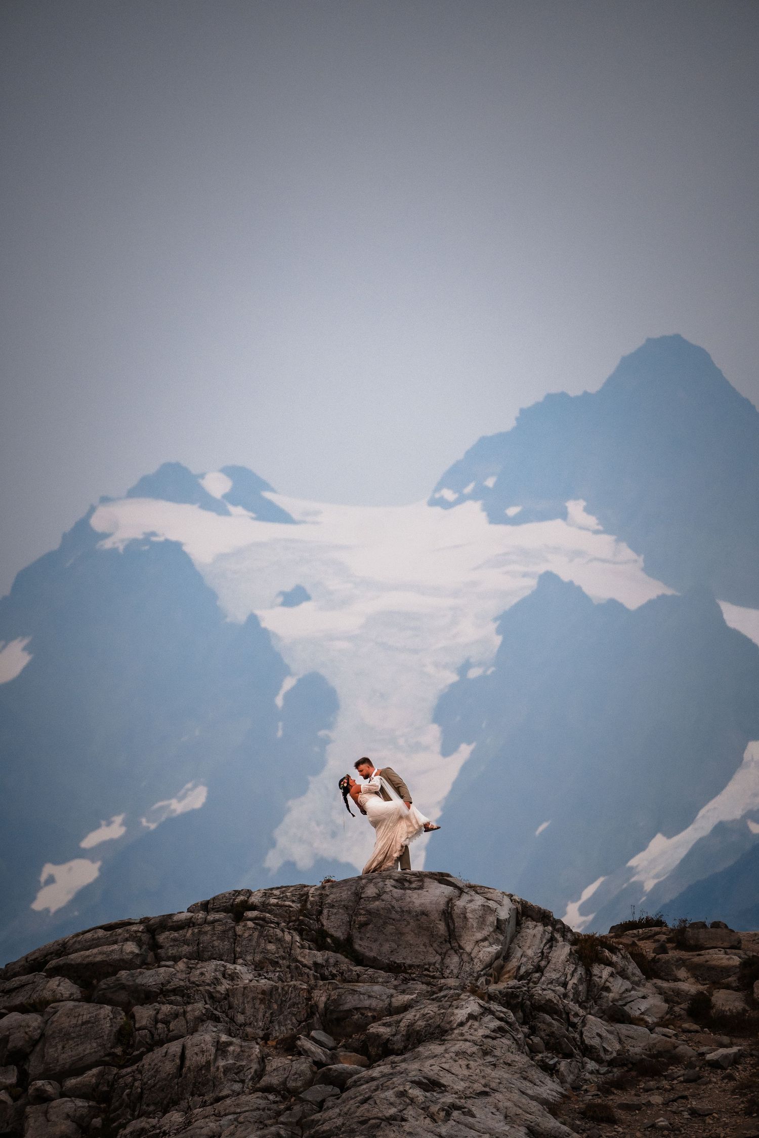 A couple poses dramatically on mountain rocks with snow-capped peaks and glaciers in the background.