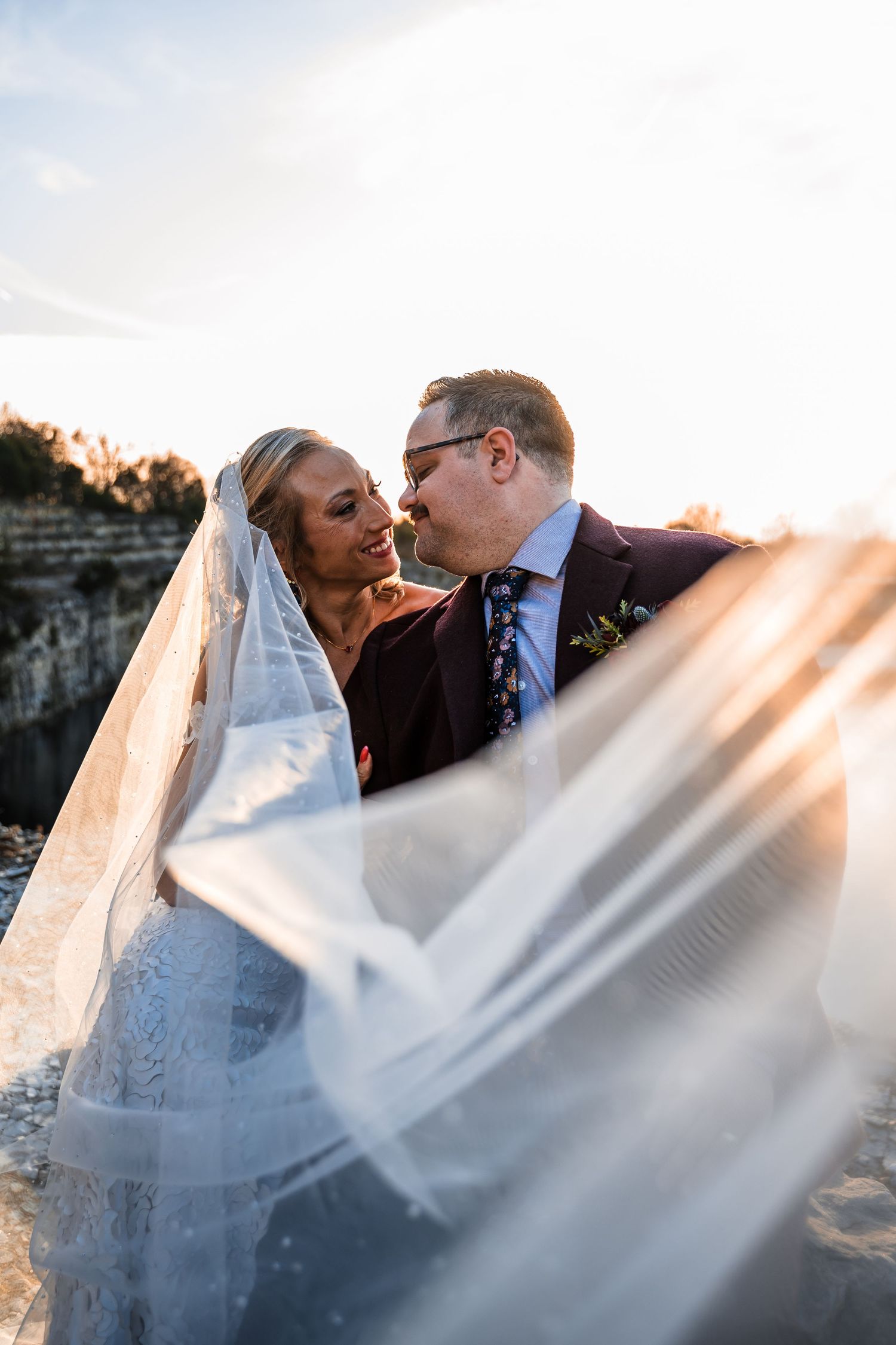 Romantic wedding couple embraces under flowing white veil during sunset photo session outdoors.