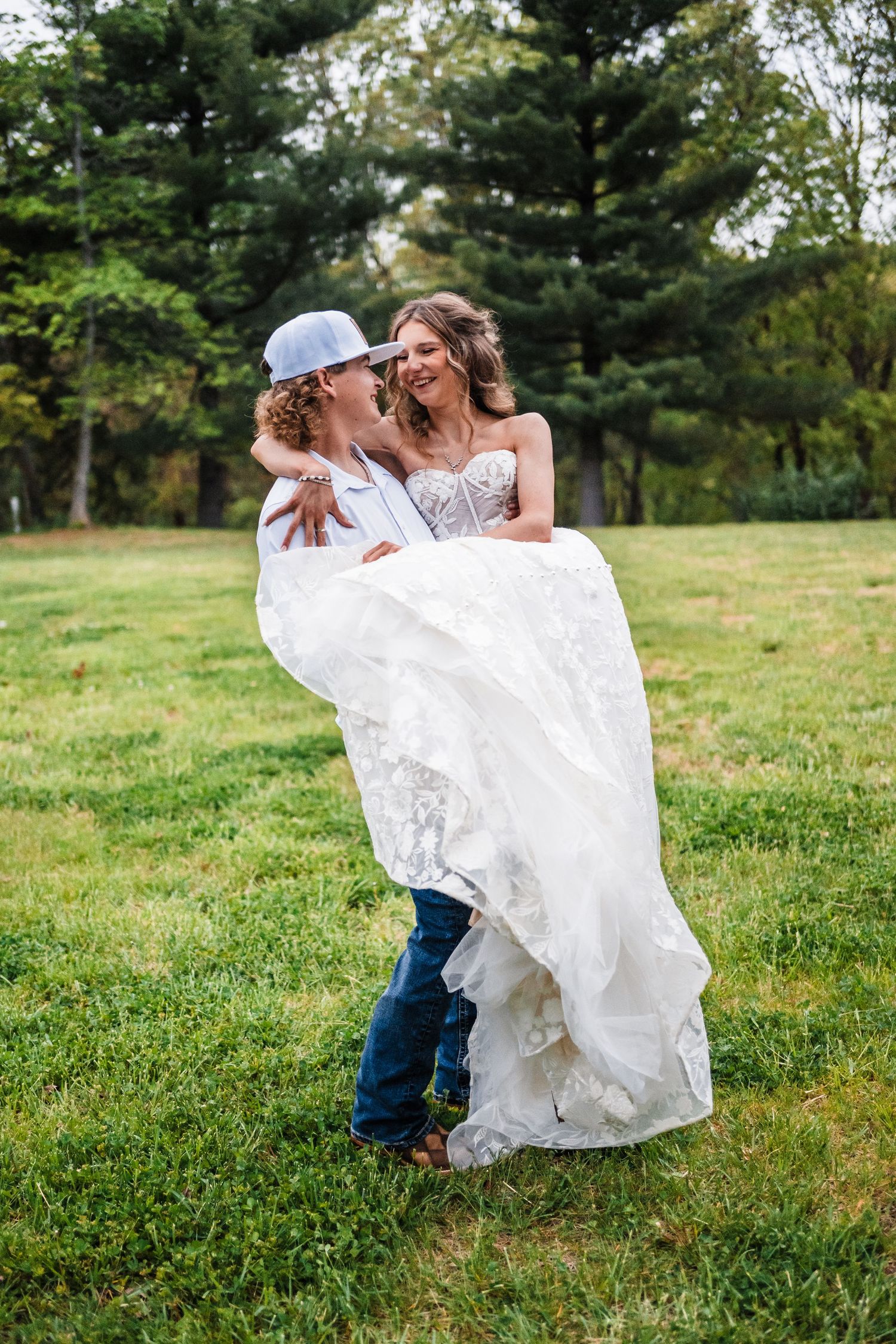 A joyful wedding couple embraces in a grassy field with pine trees in the background.