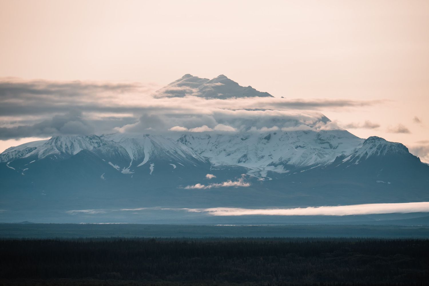 Snow-capped mountain peak rises above low-lying clouds in a moody blue landscape at dusk.