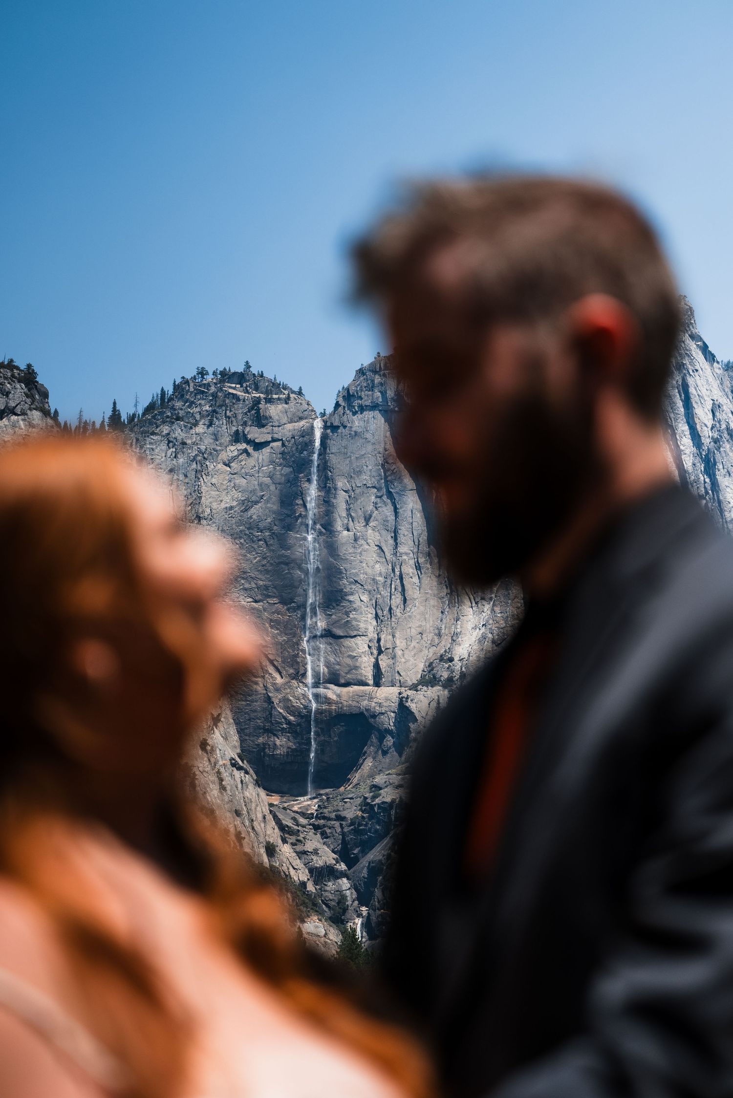 Wedding couple silhouetted against towering Yosemite Falls and blue mountain landscape.