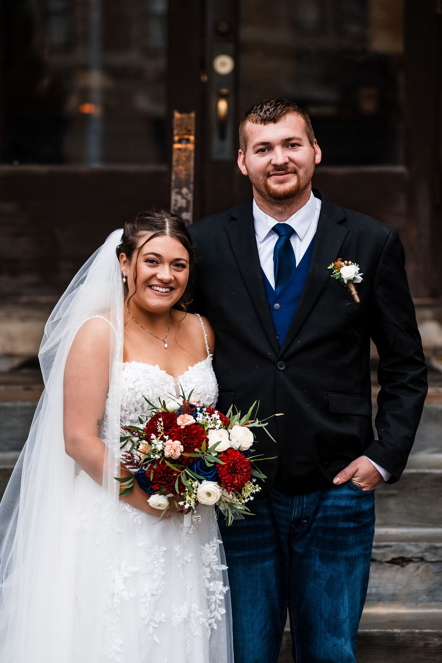 Newlywed couple in navy and white attire pose for wedding portraits with burgundy and cream floral bouquet.