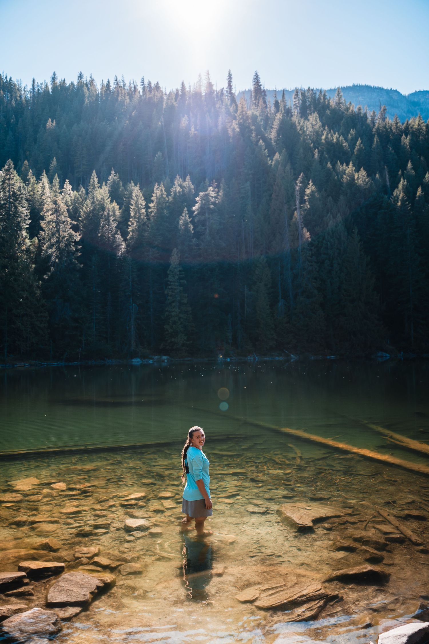 Person standing in shallow mountain lake surrounded by sunlit pine forest.