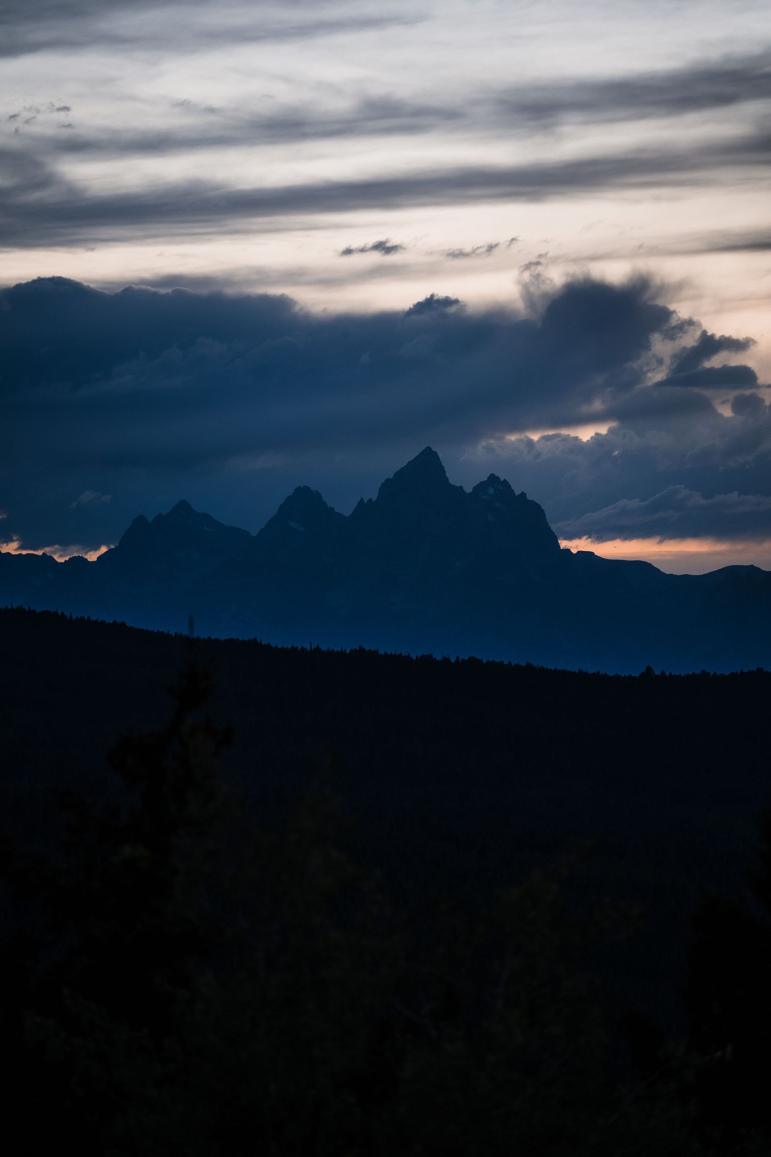 A dramatic mountain silhouette against a moody cloudy sunset sky creates a striking dark landscape panorama.