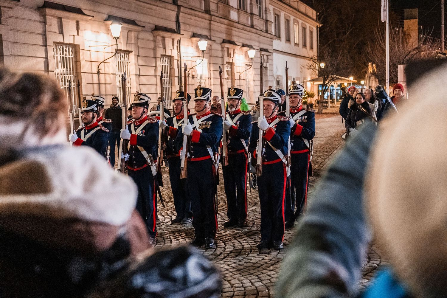 Military personnel in ceremonial uniforms march in formation during a traditional parade through historic city streets.