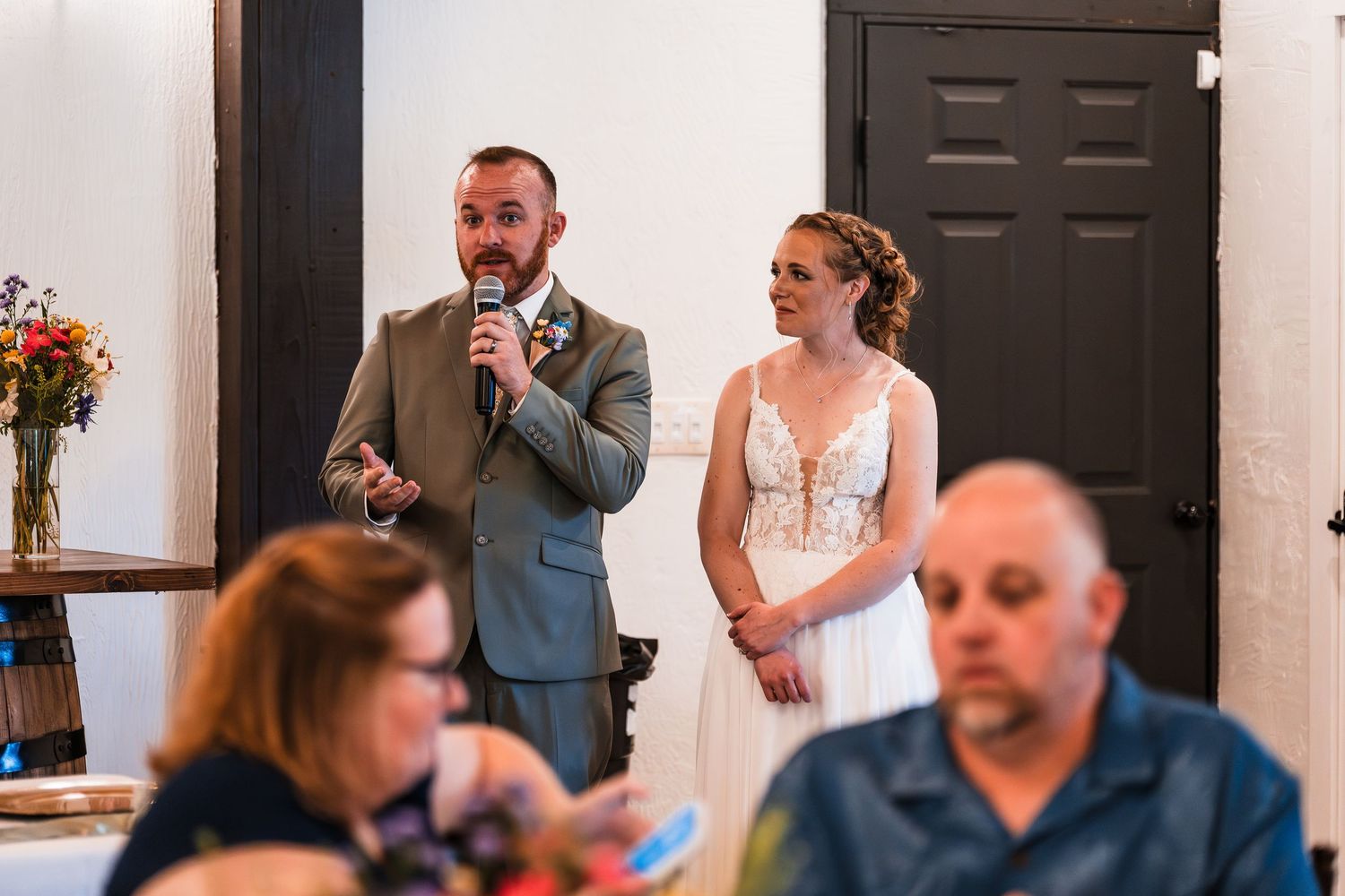 Groom speaks into microphone at wedding reception while bride in lace dress stands beside him.