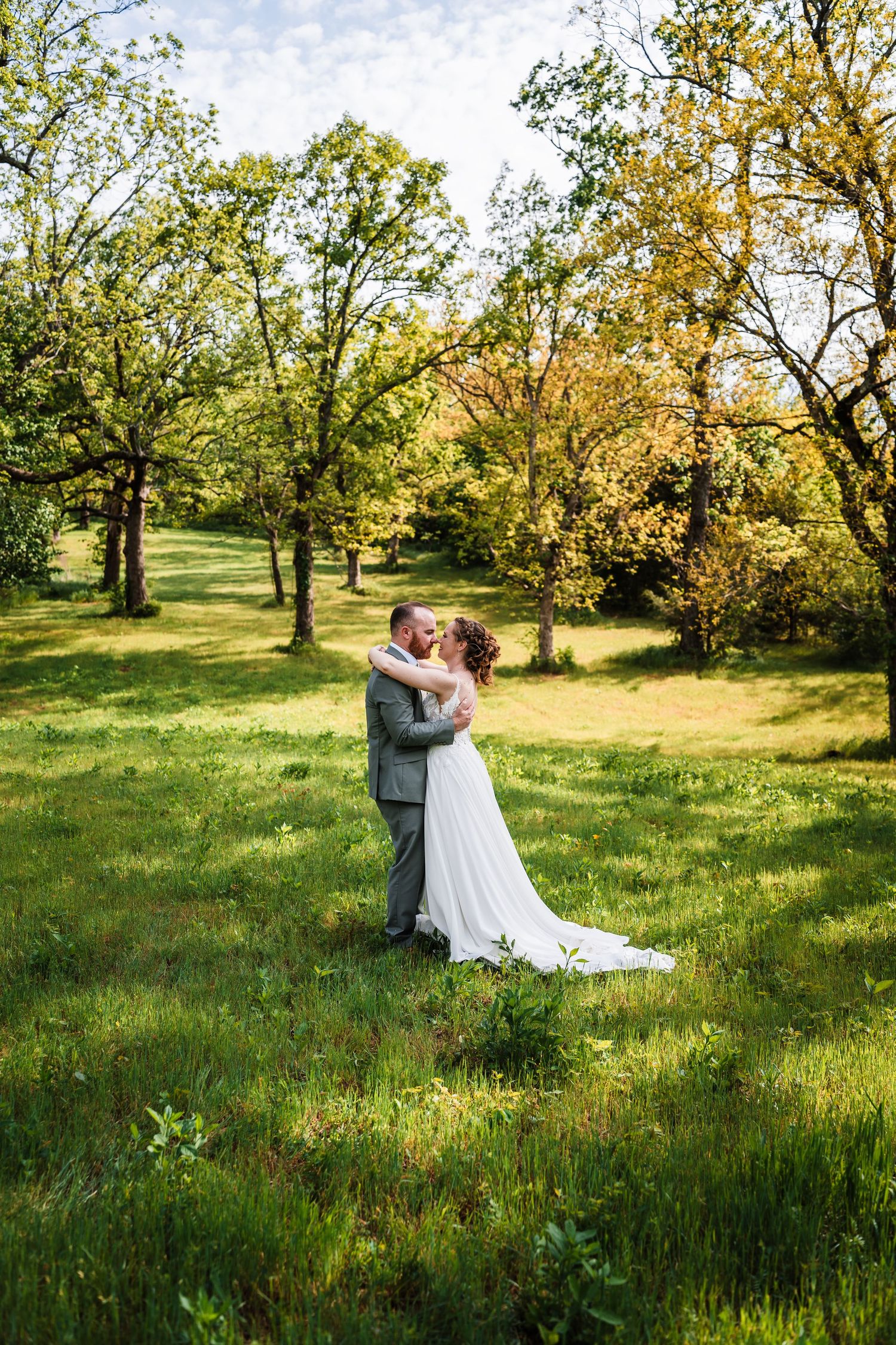 Bride and groom sharing a romantic kiss in a lush green meadow surrounded by trees.
