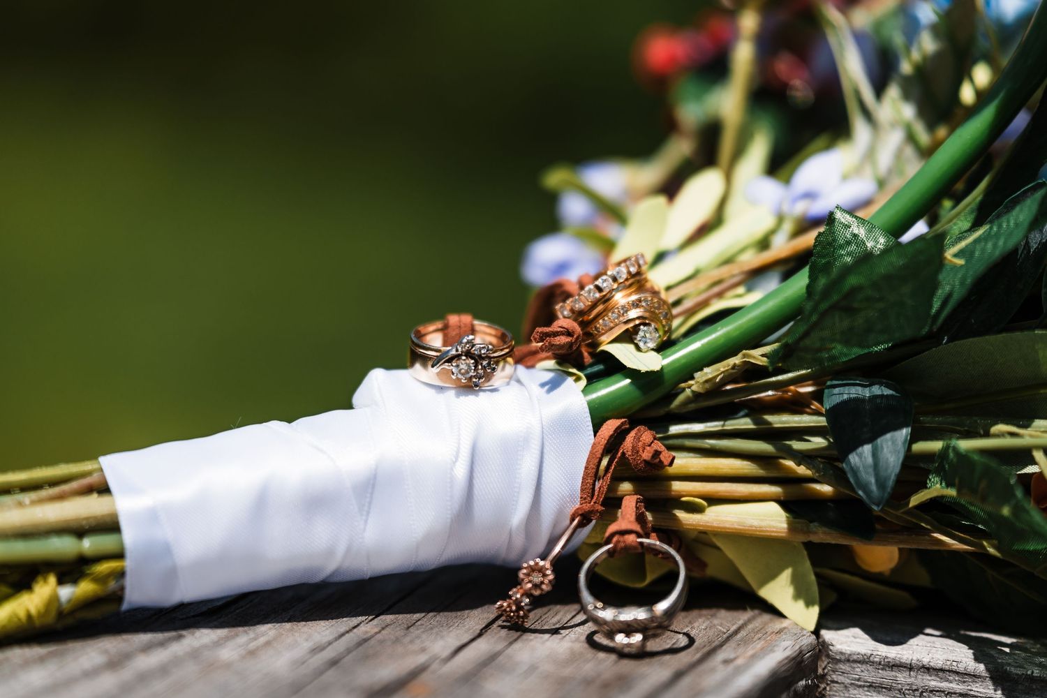 Wedding rings and a key charm rest on a white ribbon wrapped around a bridal bouquet on a rustic wooden surface.