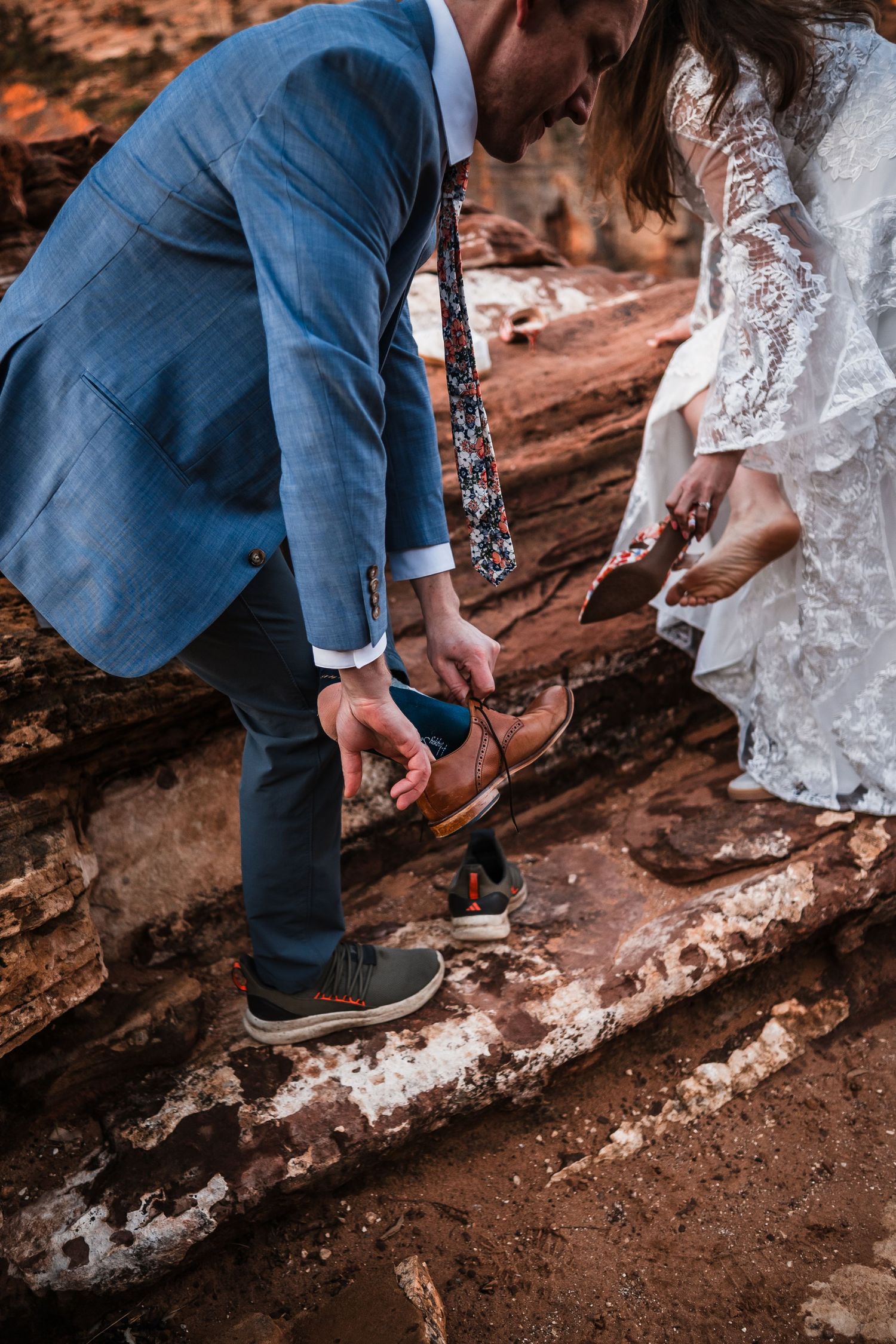 Groom in blue suit helps bride with brown boot on red rock terrain during outdoor wedding.