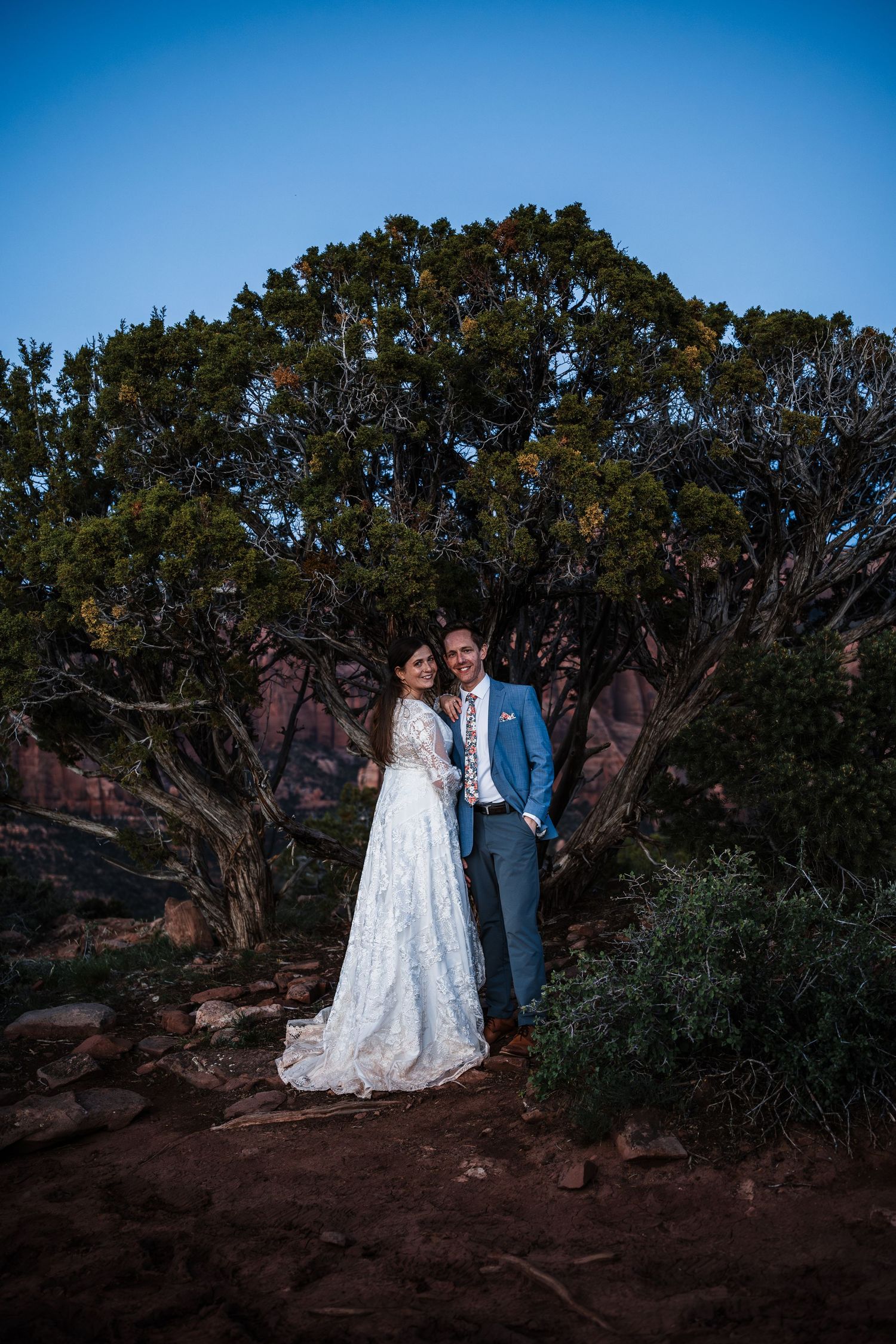Bride and groom embrace before a gnarled desert tree at dusk, red rock landscape behind them.