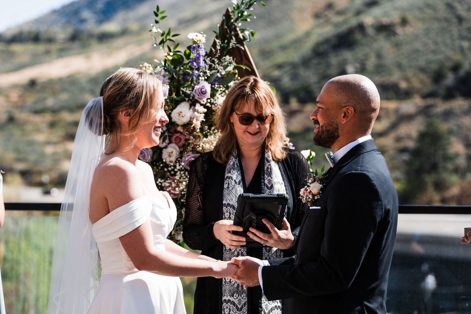 Bride and groom hold hands during wedding ceremony officiated by woman outdoors in mountains.