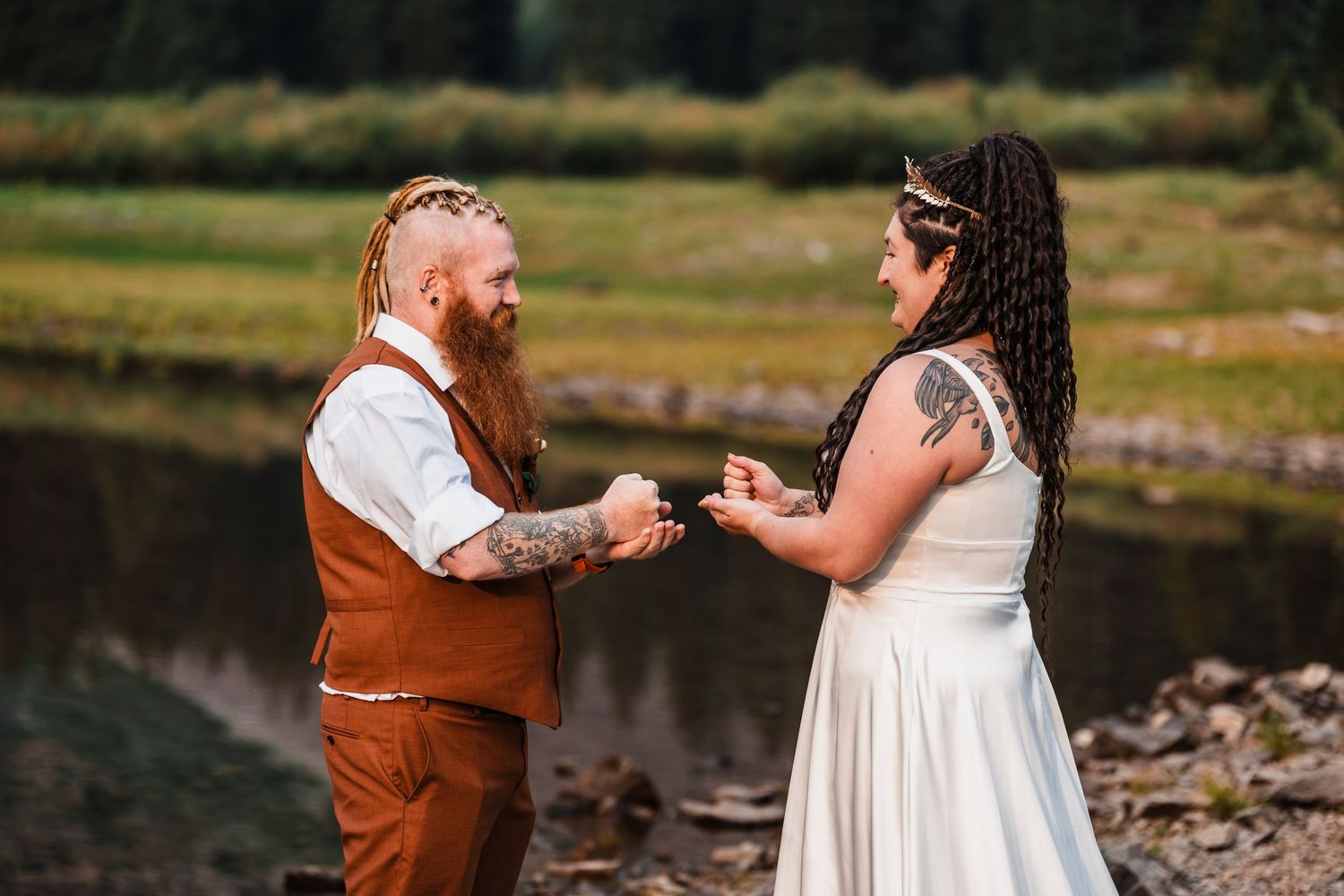 Couple exchanges vows outdoors near a mountain lake, groom in brown vest with red beard, bride in white dress with tattoos.