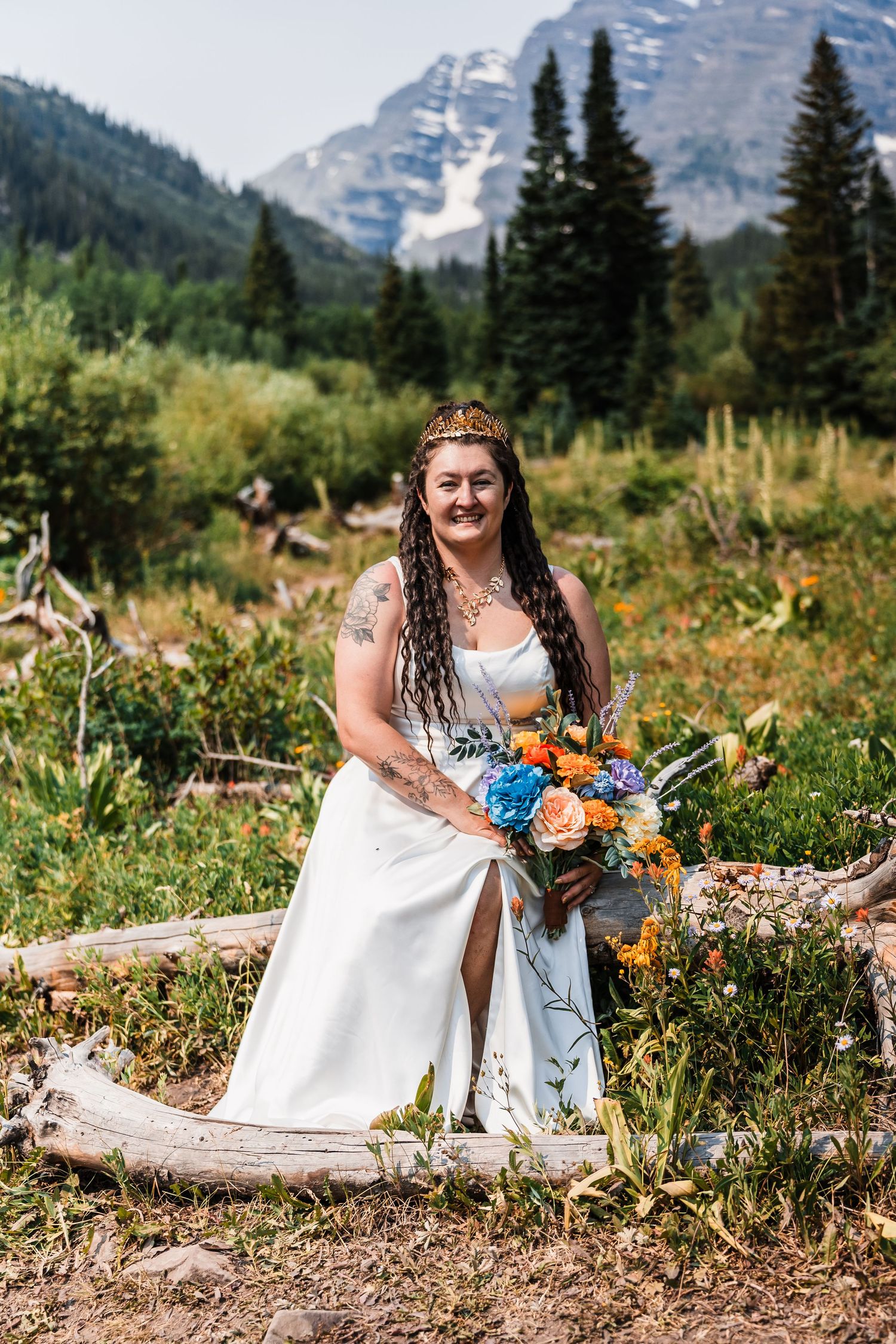 Bride with dreadlocks and gold crown sitting on log holding colorful bouquet with mountain backdrop.