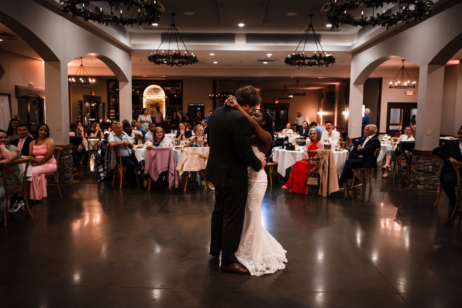 Bride and groom sharing their first dance in an elegant reception hall with seated guests watching.