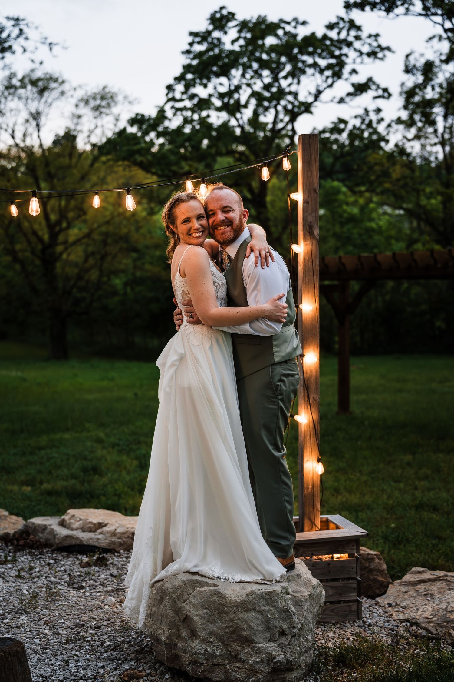 Bride and groom embracing at dusk beside rustic string lights and fire pit outdoors.