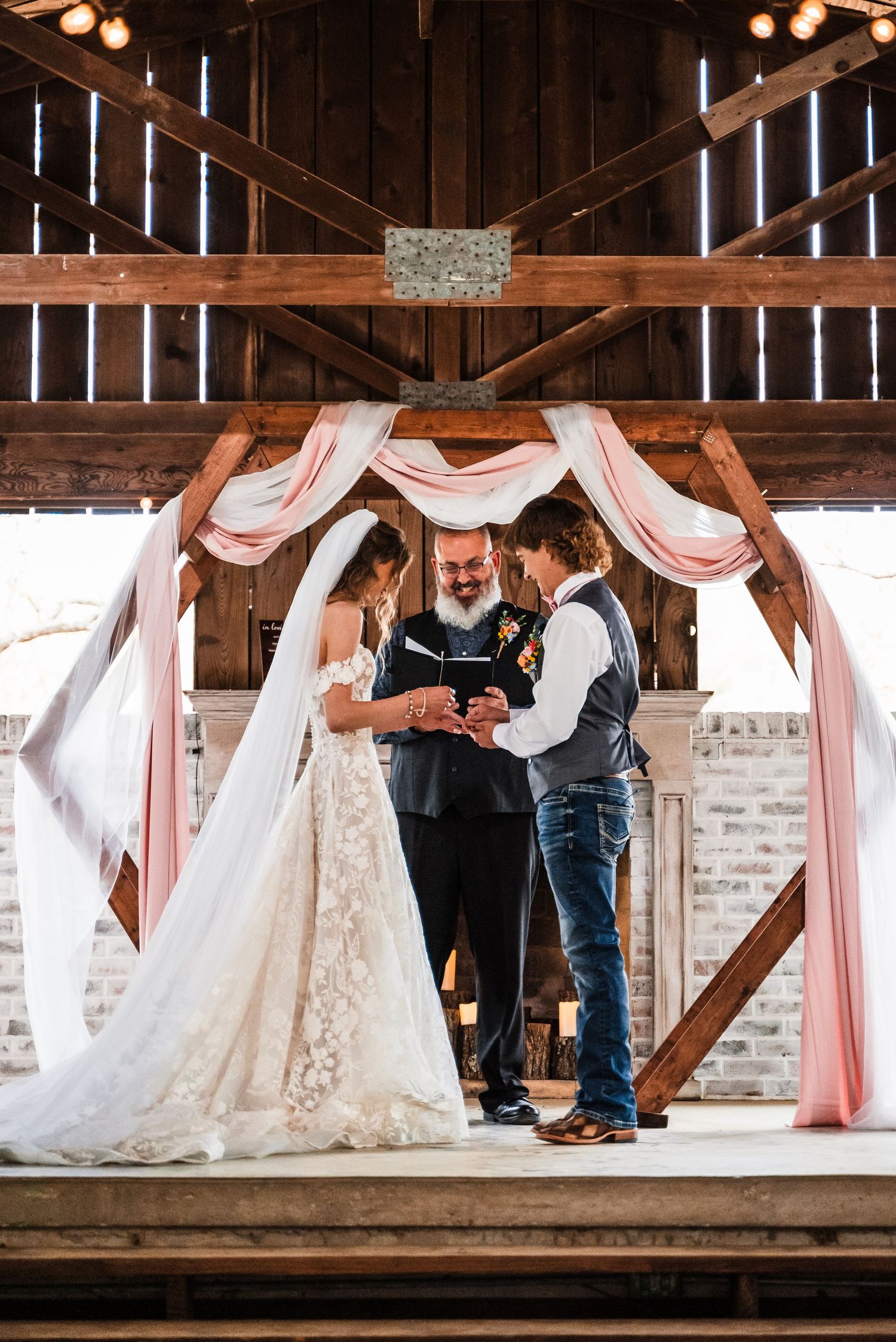 A wedding ceremony takes place under pink fabric draping in a rustic barn setting.