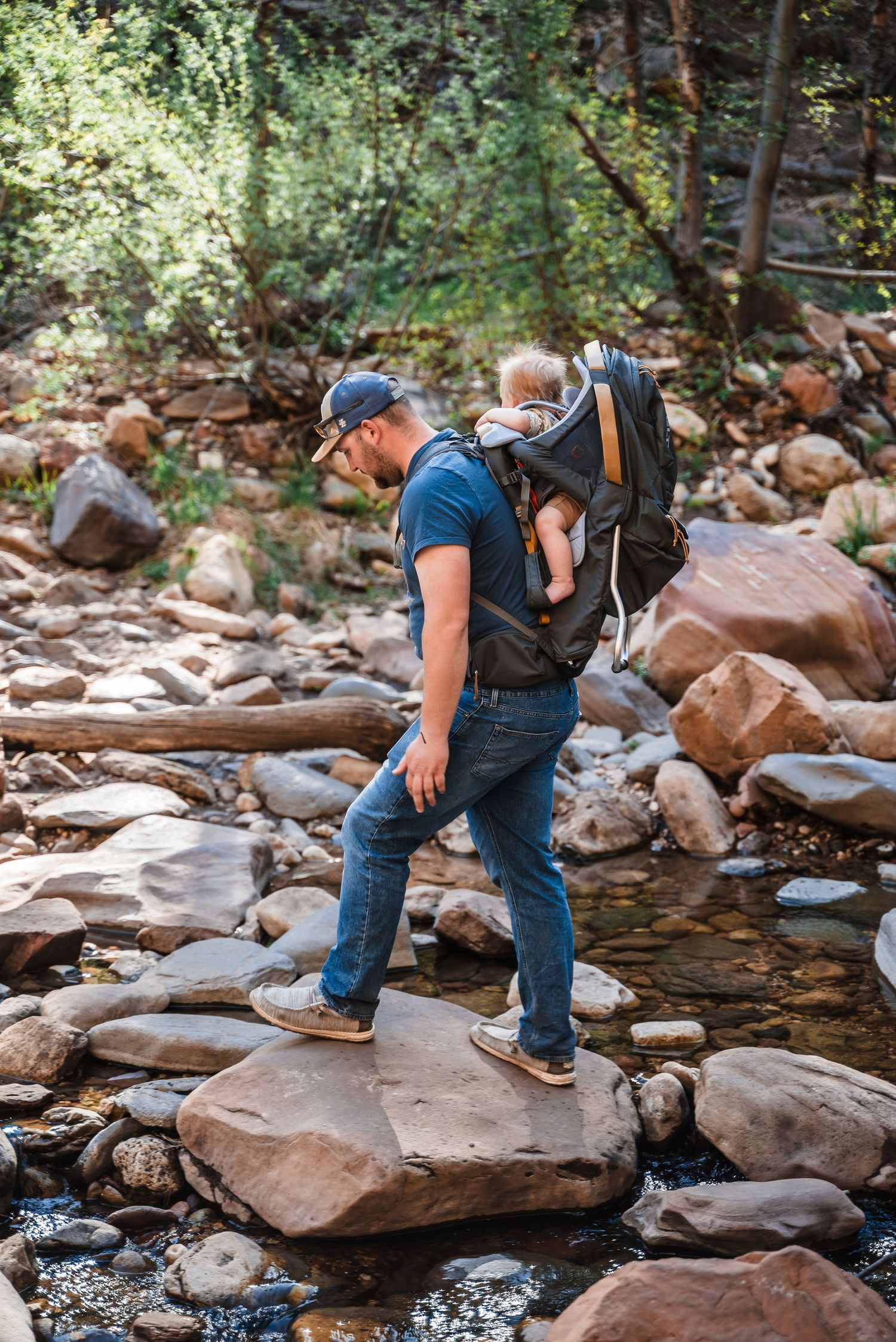 Hiker with a baby carrier backpack carefully crosses rocky stream bed in a wooded setting.