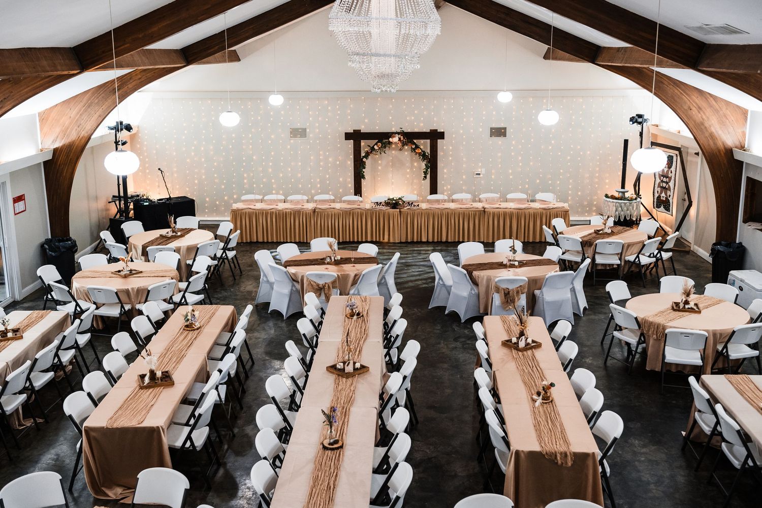 Wedding reception venue with rustic wooden beams, long banquet tables, white chairs, and elegant chandelier lighting.