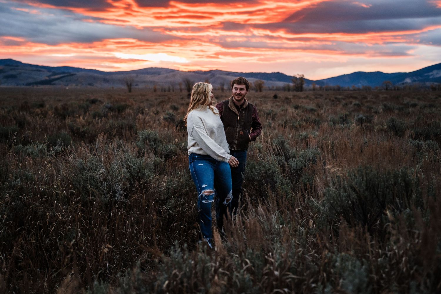Couple stands together in a desert landscape during a dramatic orange and pink sunset.
