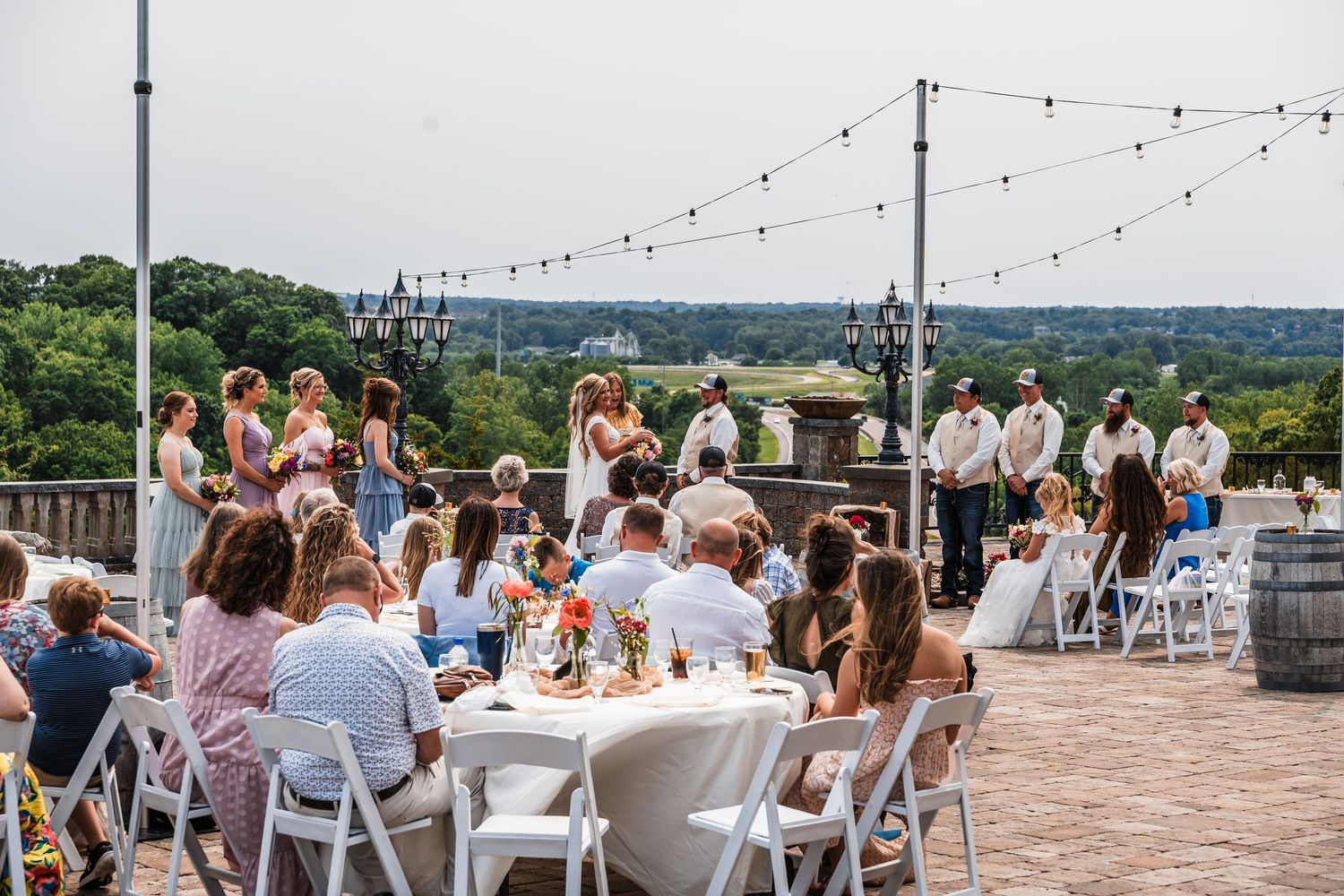 Outdoor wedding reception setup with string lights and white chairs overlooking scenic hillside views.